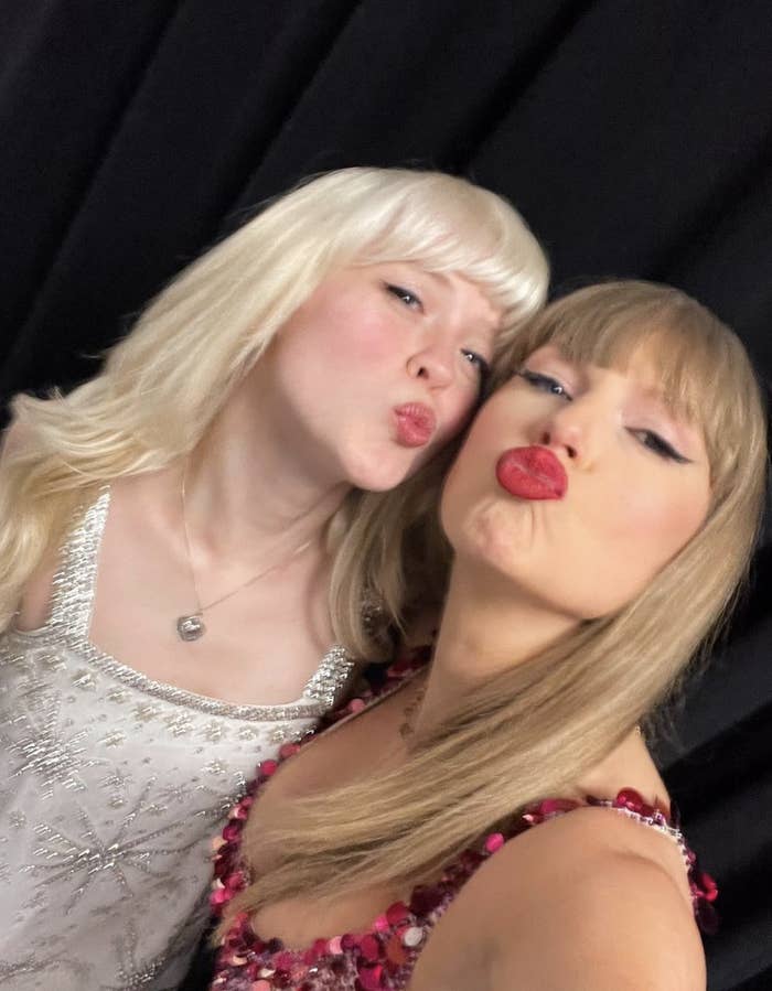 Taylor Swift and Maisie Peters pose for a selfie, making kissy faces. Taylor wears a sequin dress; Maisie is in a white dress with silver detailing