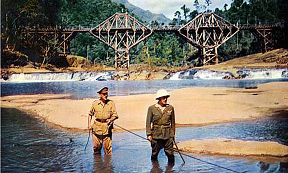 A scene from "The Bridge on the River Kwai" with two men in military attire standing in shallow water with a wooden bridge in the background