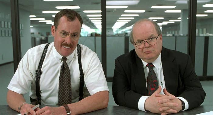 Two characters from "Office Space", one in shirt and suspenders (John C. McGinley), the other in a suit and tie (Paul Wilson), sit at a conference table