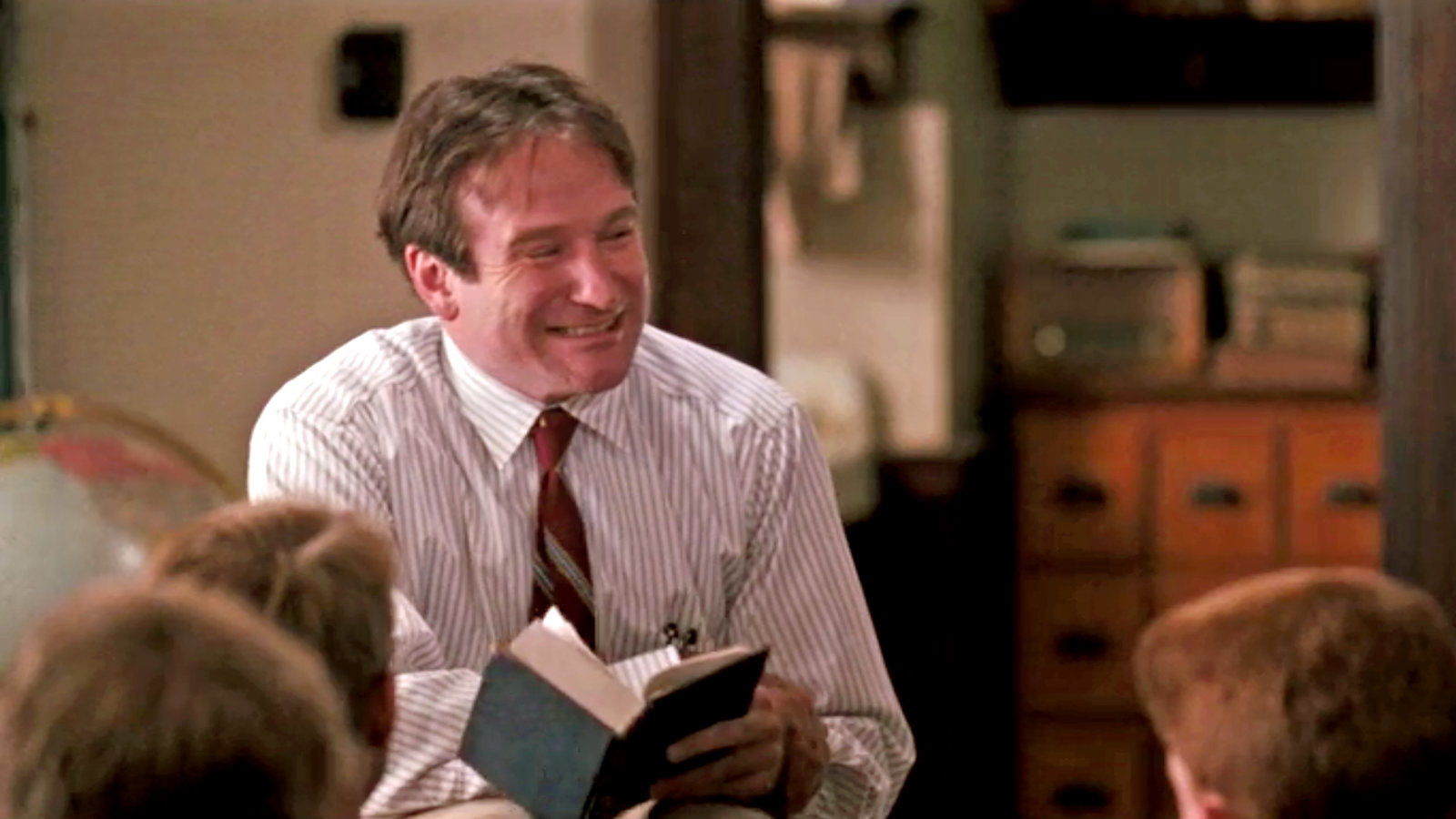 Robin Williams, smiling, holds a book in a classroom setting, engaging with students in the front row