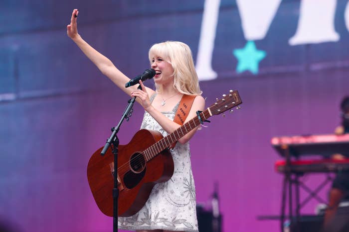 Maisie Peters in a floral dress is playing an acoustic guitar and singing into a microphone on stage, with one arm raised