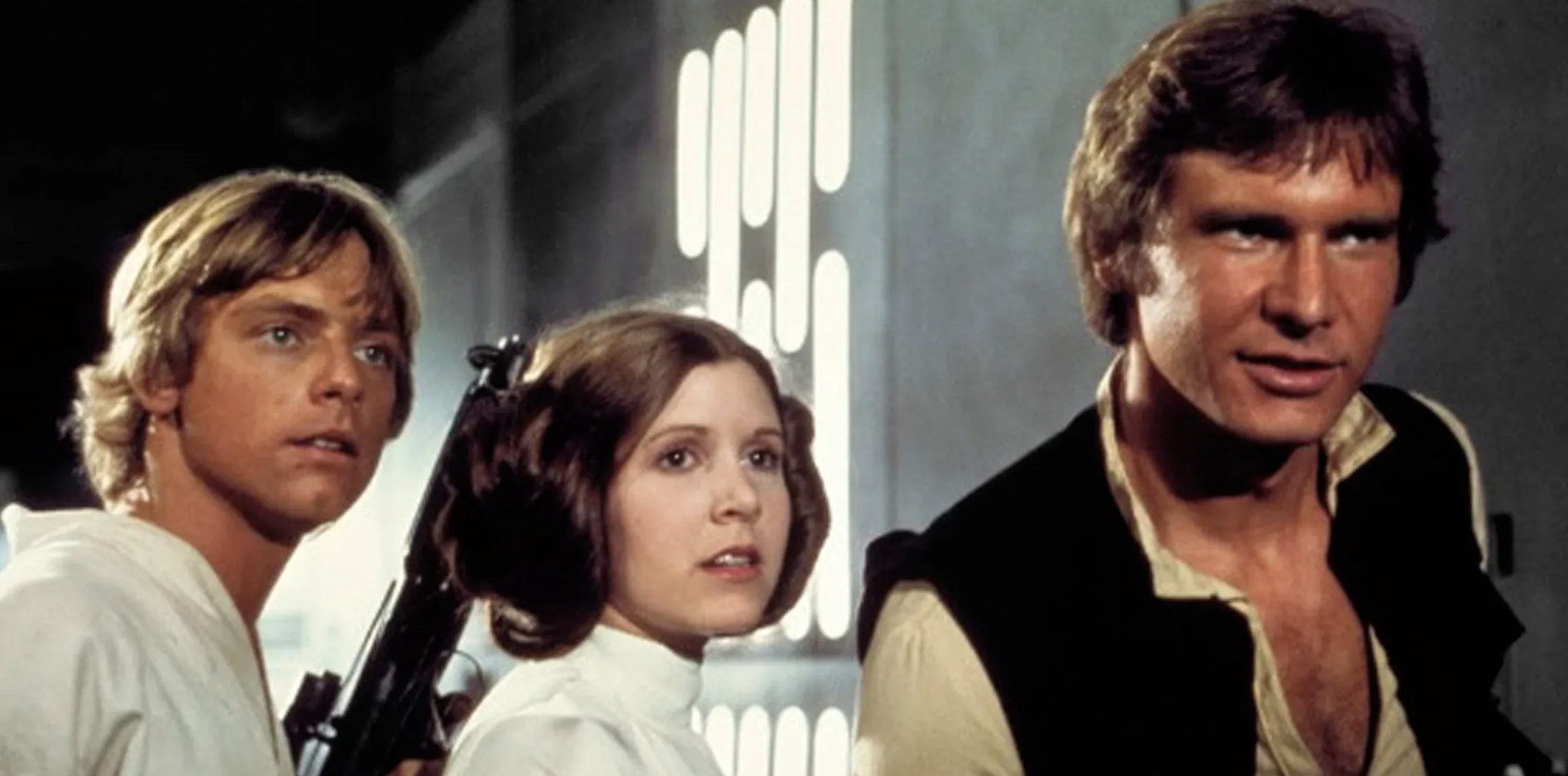 Mark Hamill, Carrie Fisher, and Harrison Ford as Star Wars characters Luke Skywalker, Princess Leia, and Han Solo, standing together and looking off-screen