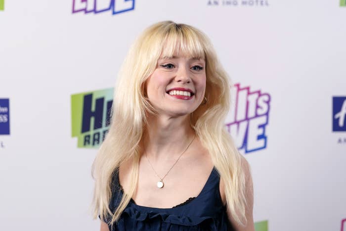 Maisie Peters with long blonde hair, wearing a sleeveless navy top, stands smiling at a promotional event backdrop with &quot;Hits Live&quot; text