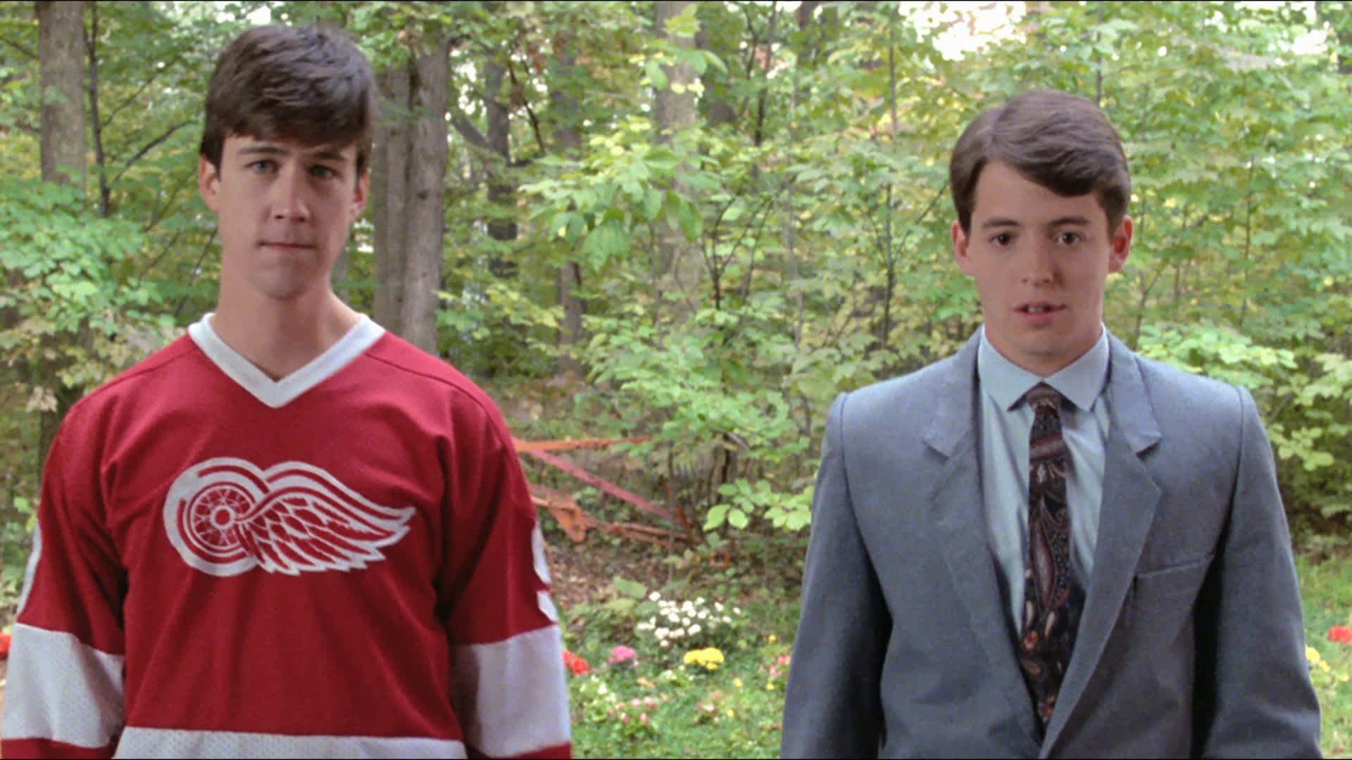 Alan Ruck in a hockey jersey and Matthew Broderick in a suit stand outdoors with trees and flowers in the background