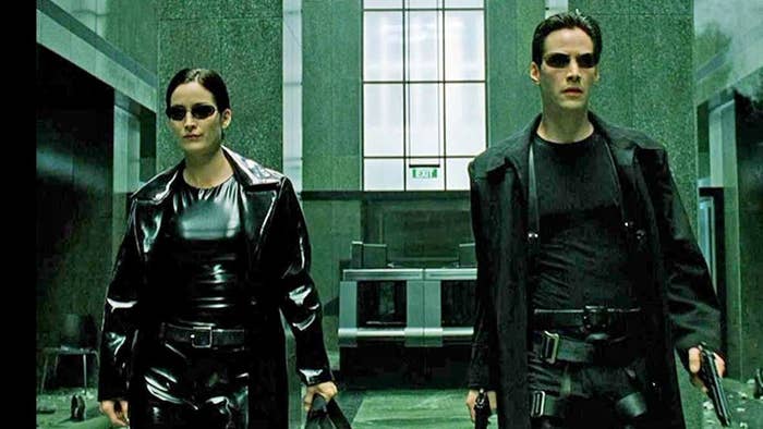 Carrie-Anne Moss and Keanu Reeves in The Matrix, wearing black leather outfits and sunglasses, walking through a building with weapons