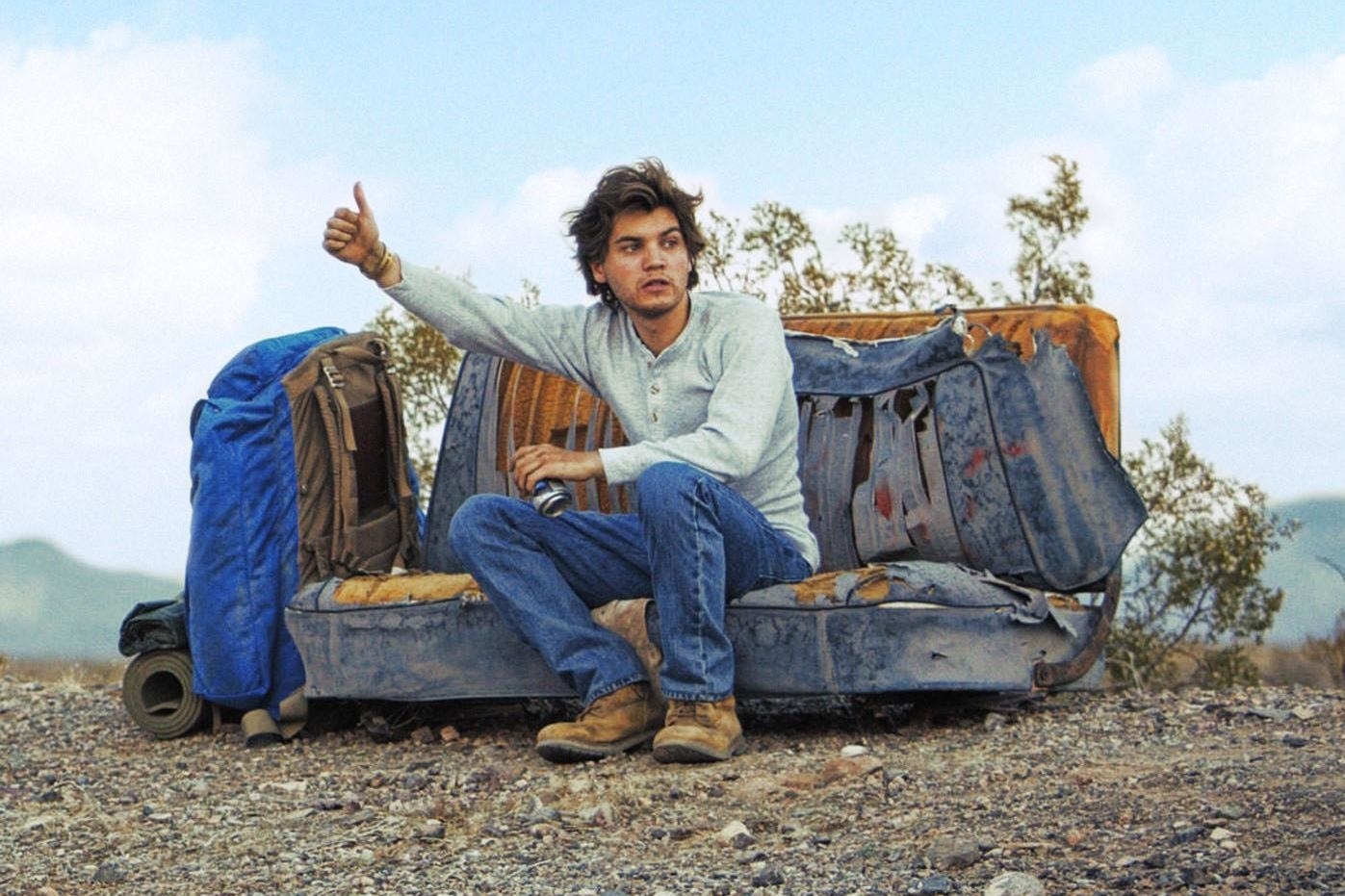 Emile Hirsch sits on a worn-out couch in a desert, hitchhiking with his thumb up, surrounded by camping gear