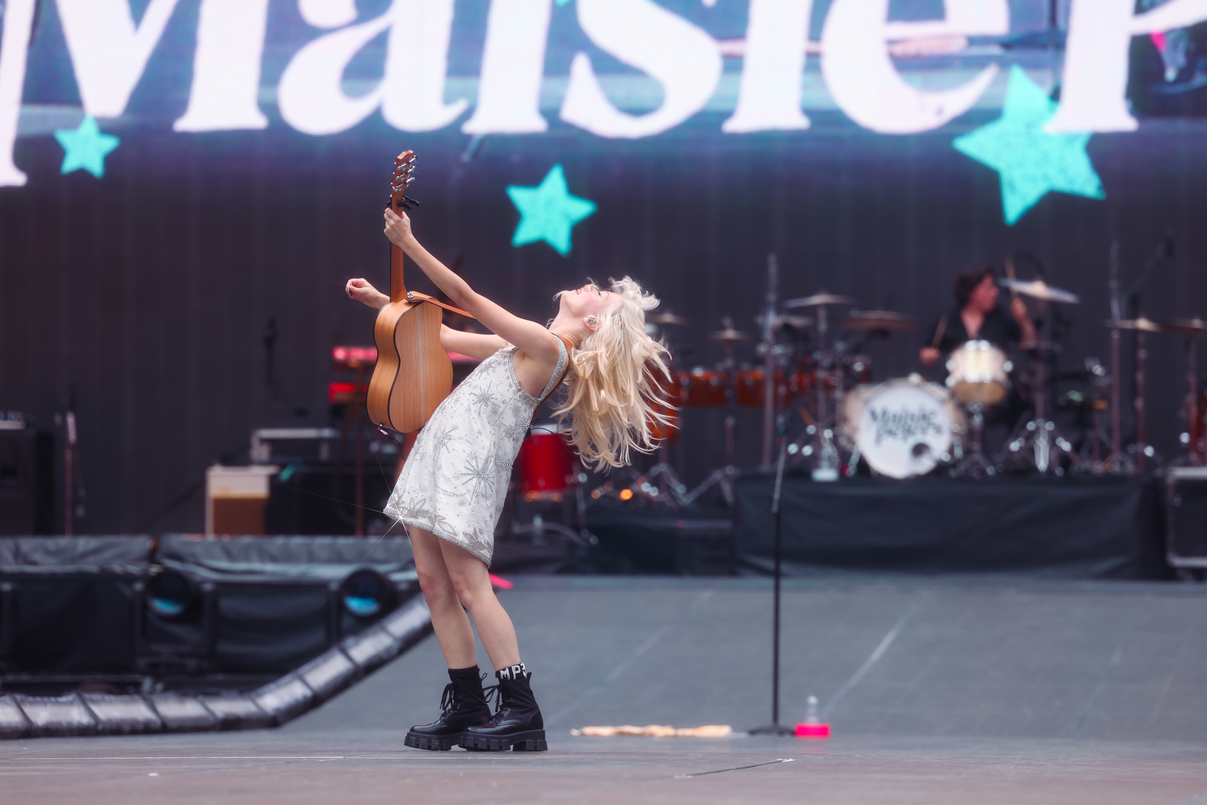 Maisie Peters plays guitar and leans back passionately on stage with a drummer visible in the background