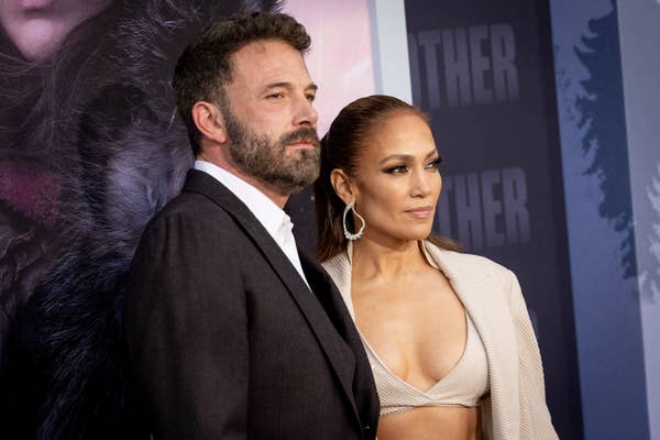 Ben Affleck and Jennifer Lopez pose at a formal event. Ben wears a suit, and Jennifer wears a stylish beige outfit with a bralette and blazer