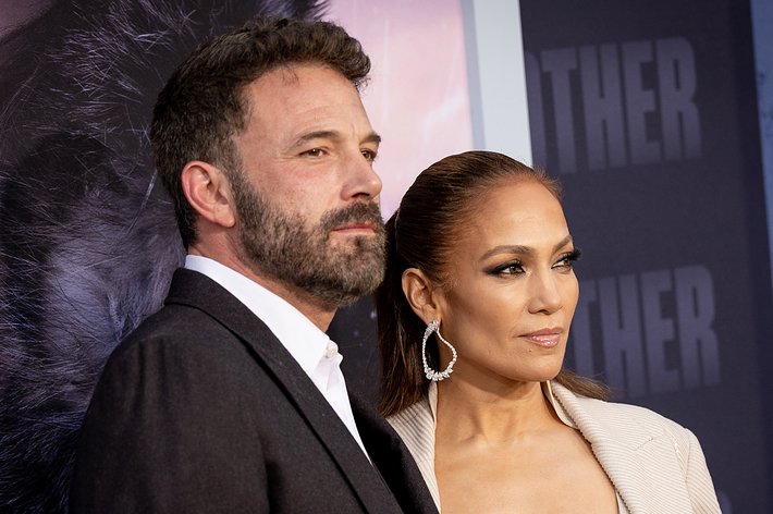 Ben Affleck and Jennifer Lopez pose at a formal event. Ben wears a suit, and Jennifer wears a stylish beige outfit with a bralette and blazer