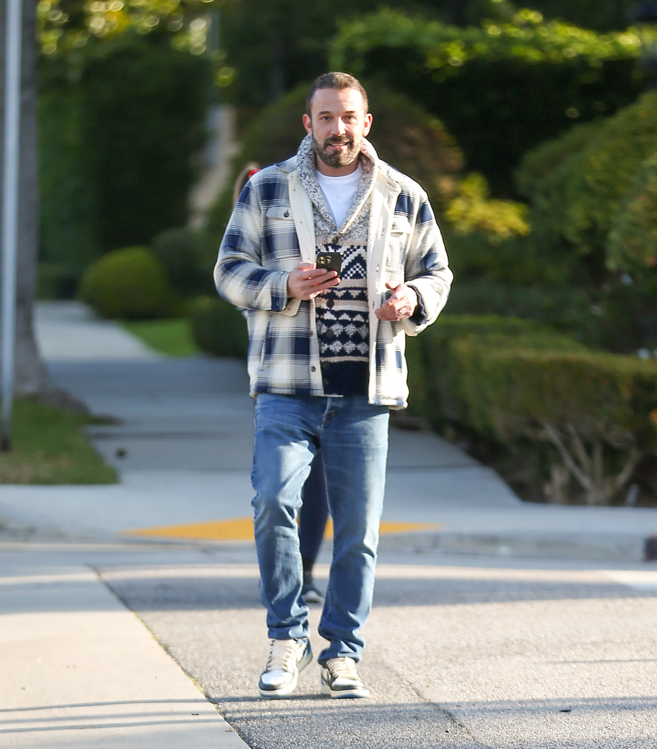 Ben Affleck walking while holding a phone, wearing a plaid jacket over a patterned sweater, jeans, and sneakers