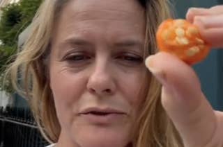 Alicia Silverstone holding up and showing a small orange fruit with seeds, standing outdoors, wearing a white blouse