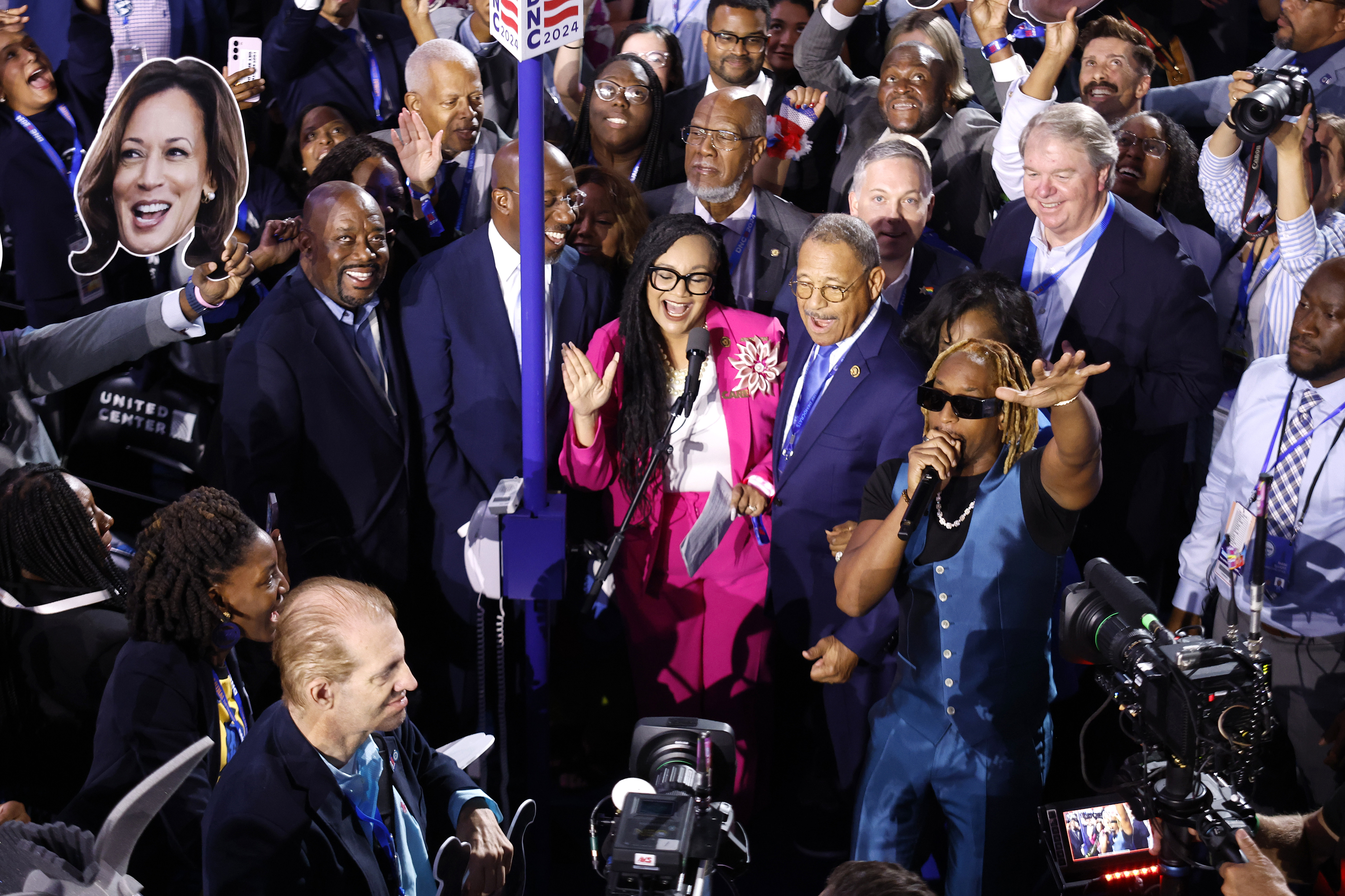 Kamala Harris cardboard cutout among a diverse group of smiling people at an event. One person speaks into a microphone; others wear business or casual attire