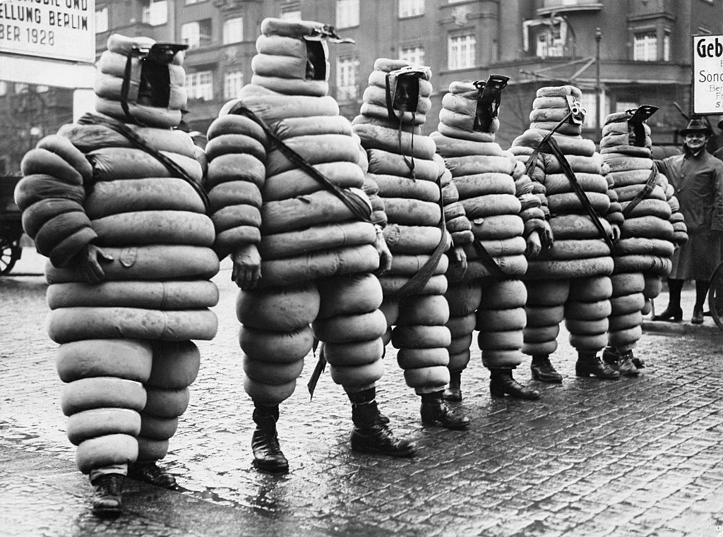 Five people in Michelin Man costumes standing in a line on a cobblestone street, circa 1920s. They appear to be participating in a promotional event