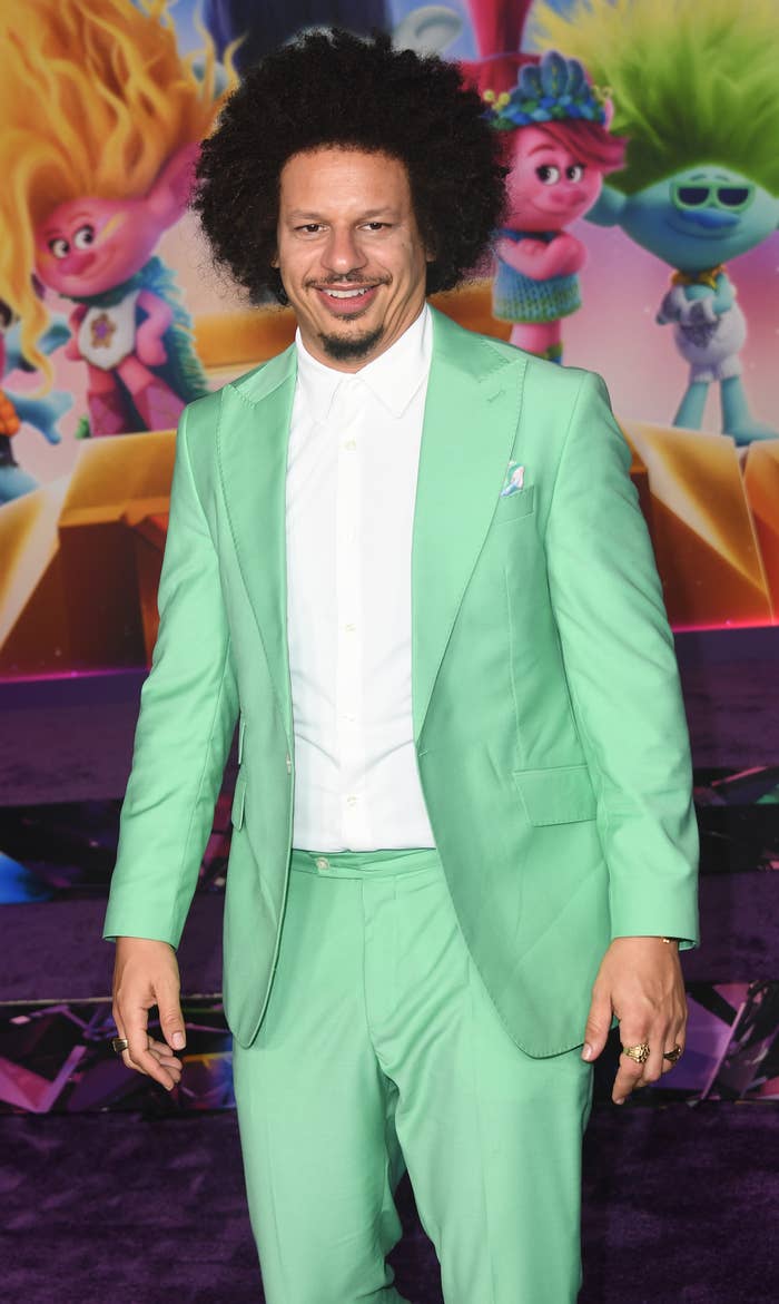 Eric André in a light green suit and white shirt at a movie premiere with Trolls characters in the background