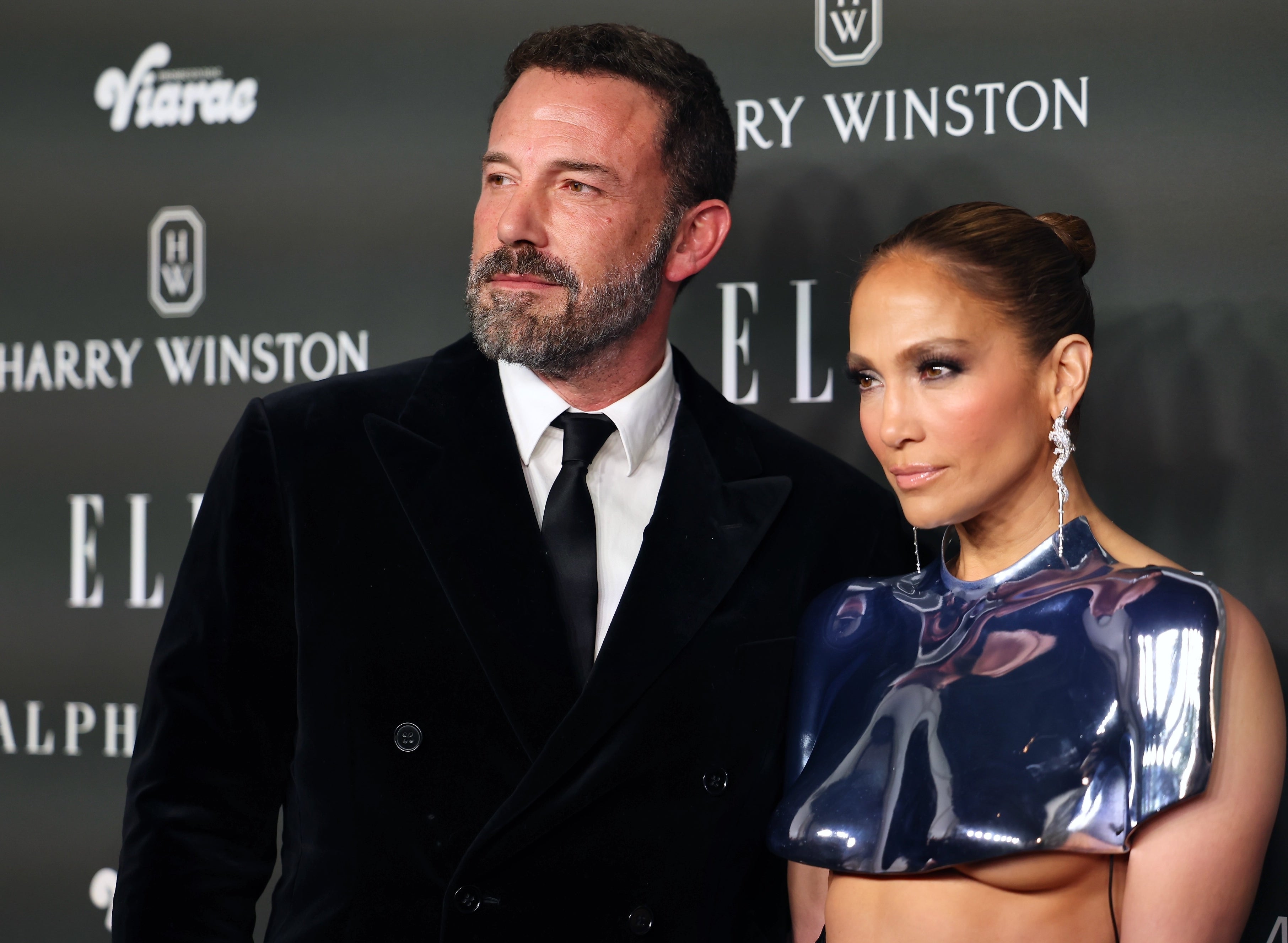 Ben Affleck in a black velvet suit and Jennifer Lopez in a high-neck, metallic cropped top at an event with logos behind them