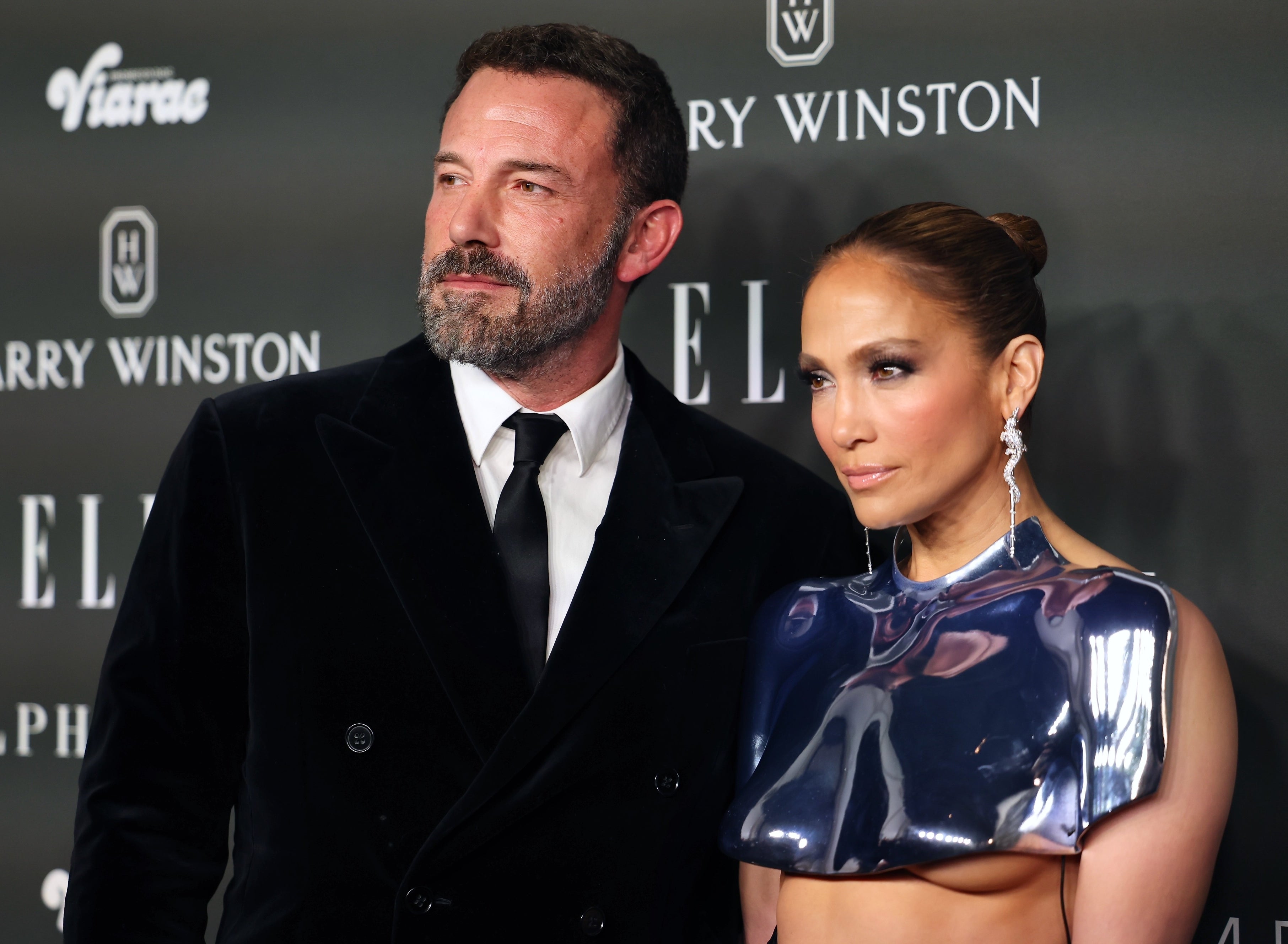 Ben Affleck in a black velvet suit and Jennifer Lopez in a high-neck, metallic cropped top at an event with logos behind them