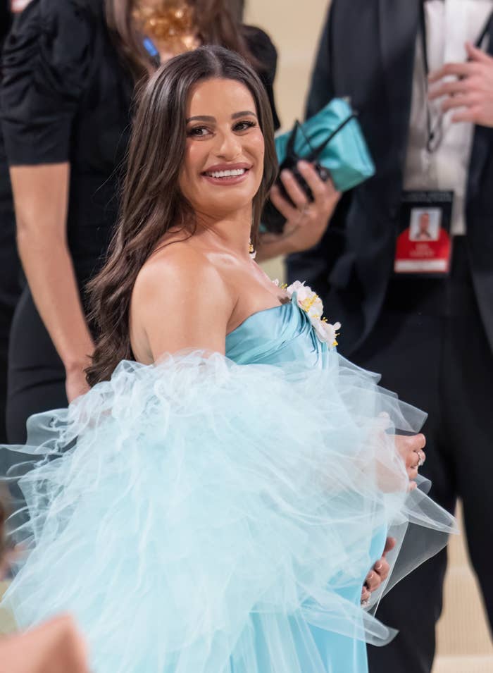 Lea Michele smiles while posing on a red carpet in an elegant strapless gown surrounded by people in formal attire