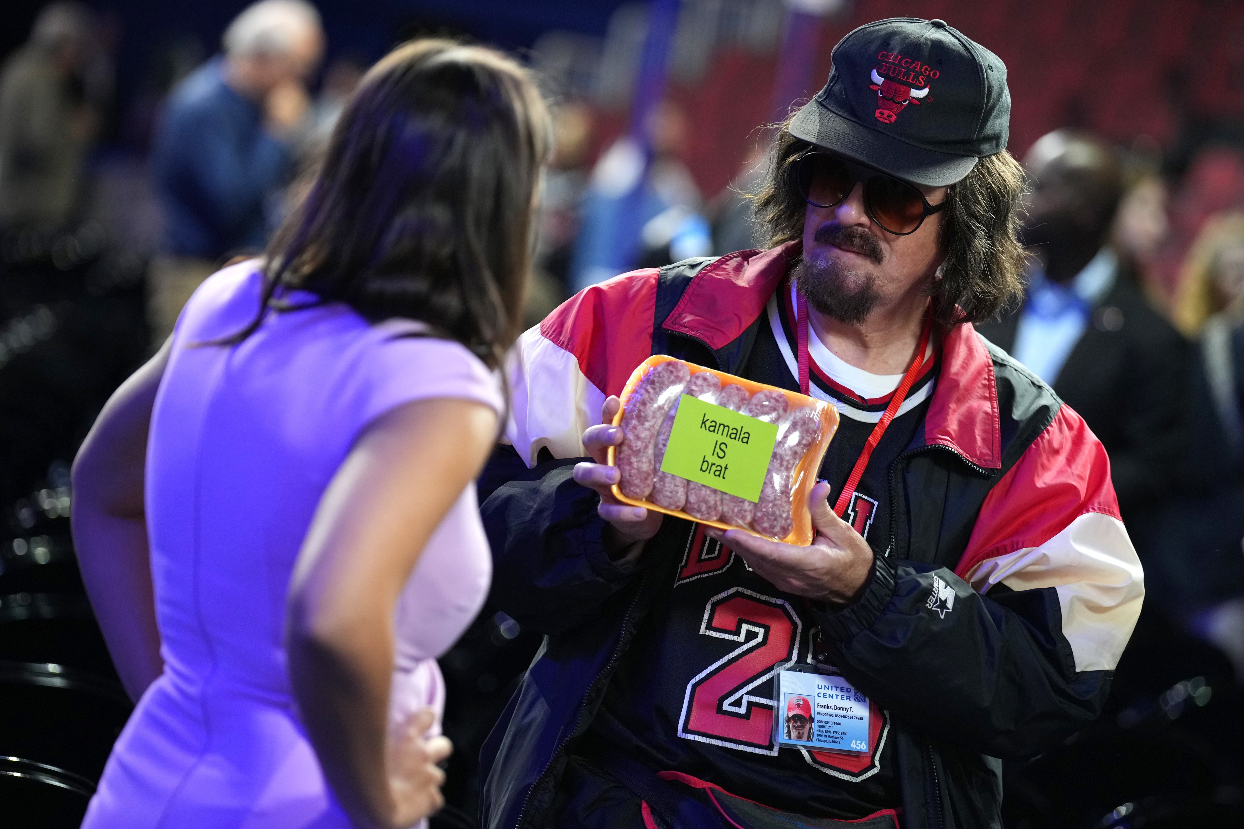 A man in a sports jacket and baseball cap holds a package with a yellow note reading "kamala is a brat" while talking to a woman