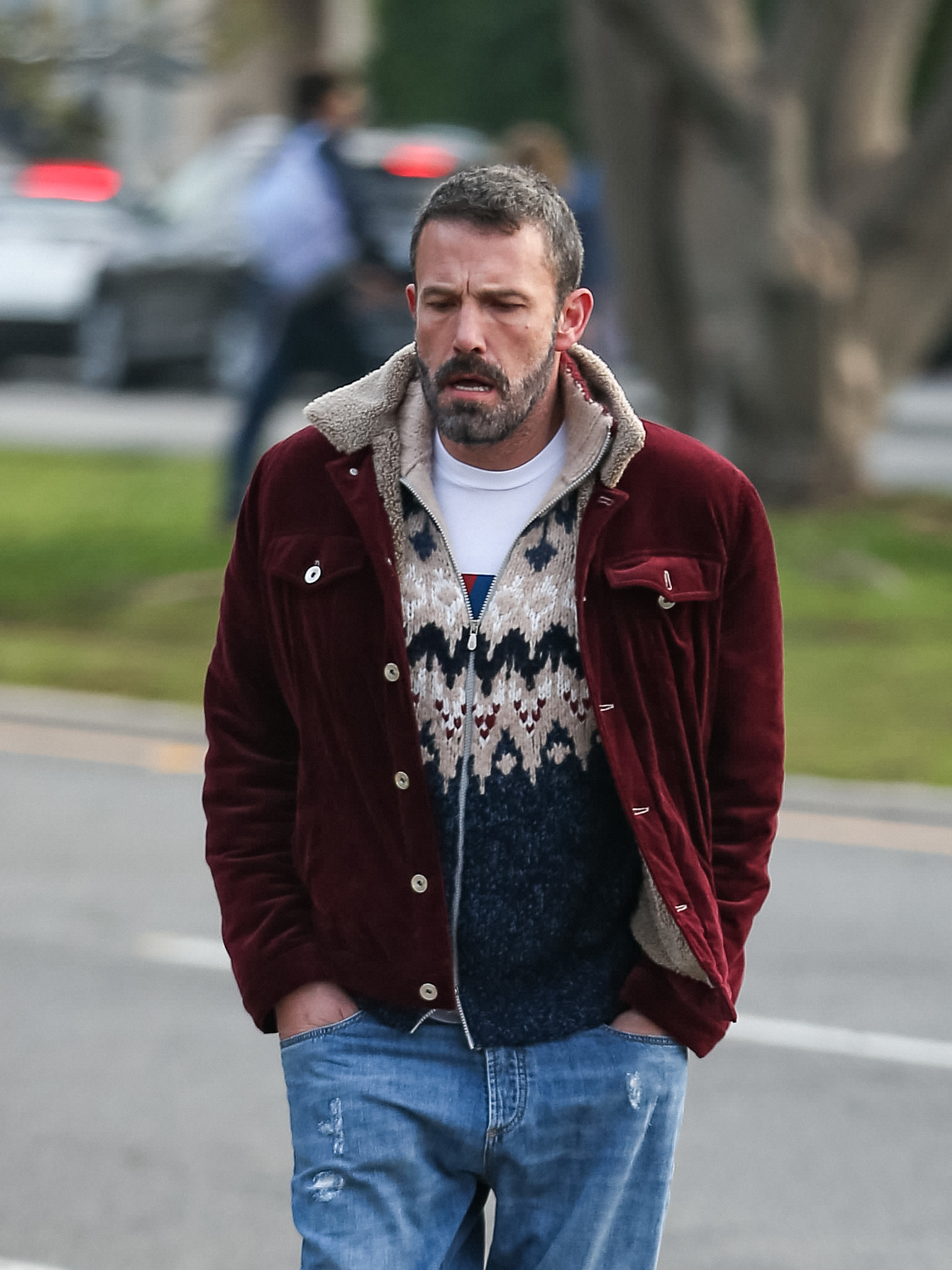 Ben Affleck walks outdoors wearing a casual outfit with a jacket over a patterned sweater and blue jeans