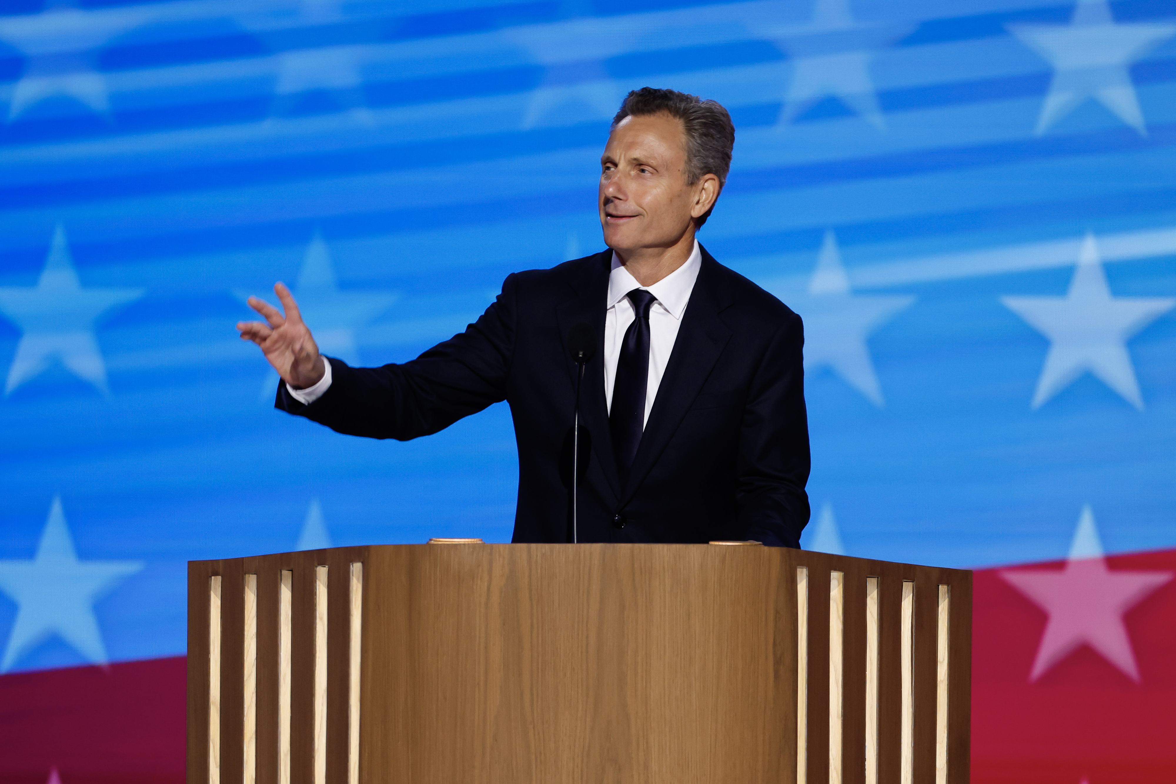 Tony in a dark suit speaks at a podium in front of a backdrop featuring stars and stripes