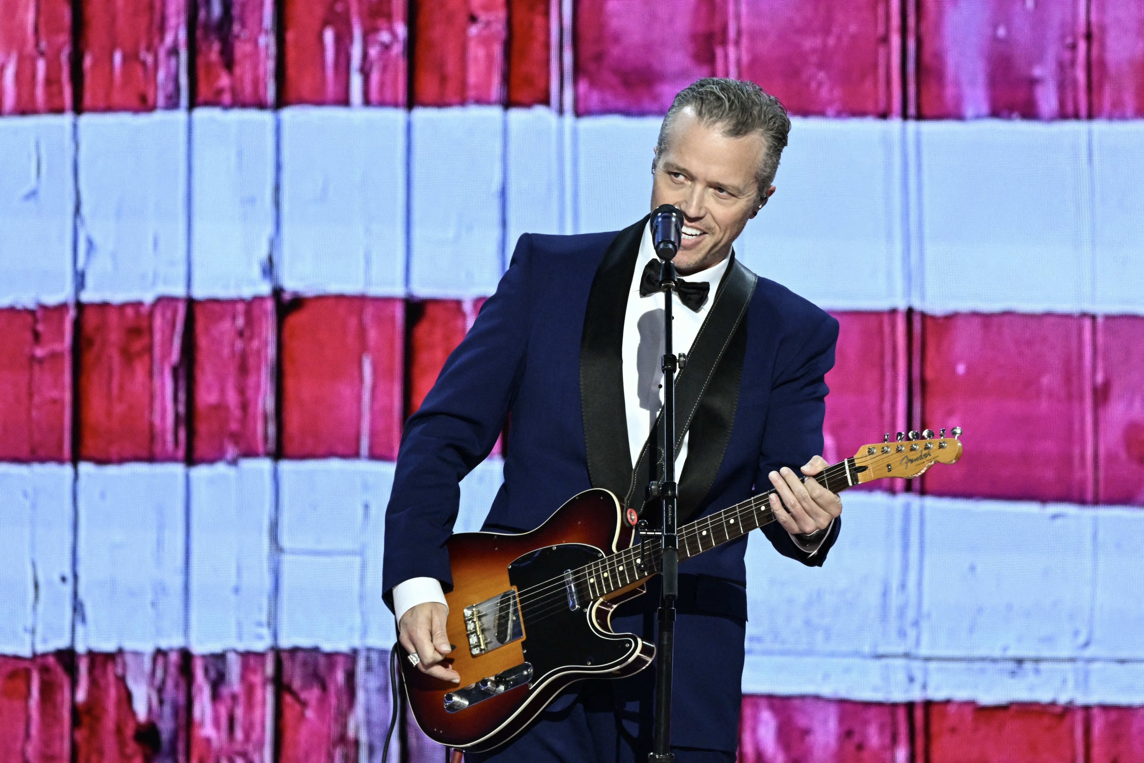 Jason Isbell, wearing a dark suit and black bow tie, performs on stage with an electric guitar against a backdrop of horizontal stripes