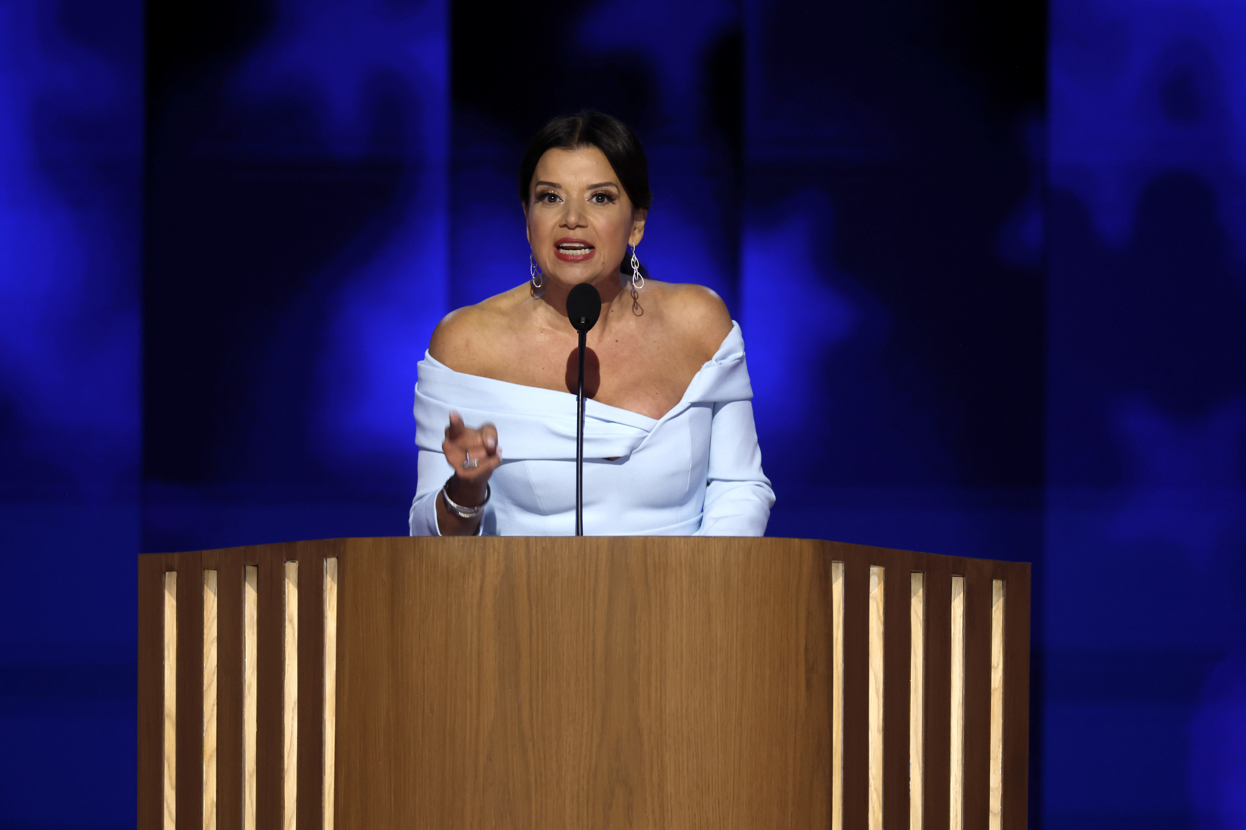 A woman stands behind a wooden podium, speaking into a microphone at an event. She is wearing an off-the-shoulder dress