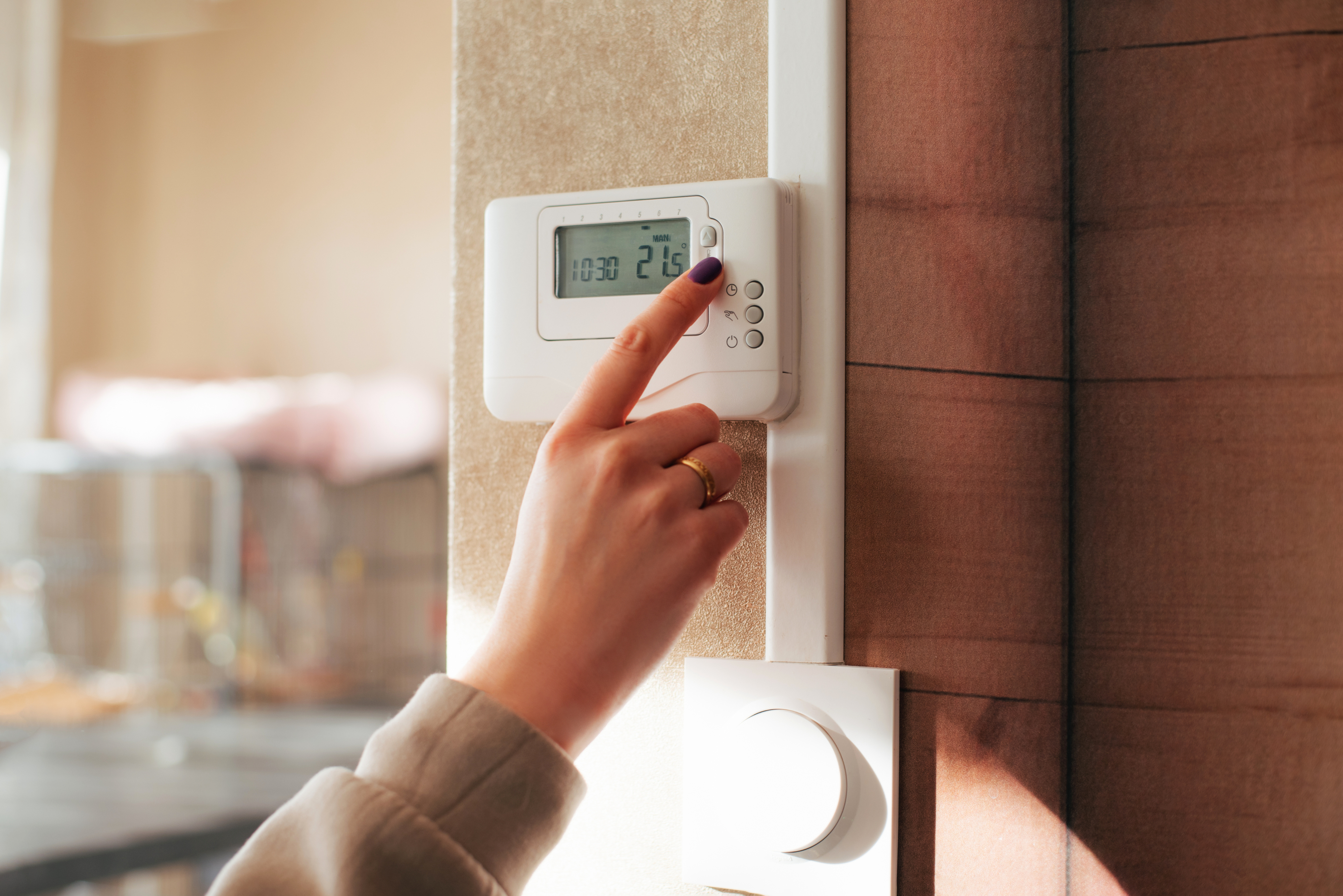 A hand with a ring adjusts a home thermostat on the wall, setting it to 21 degrees Celsius