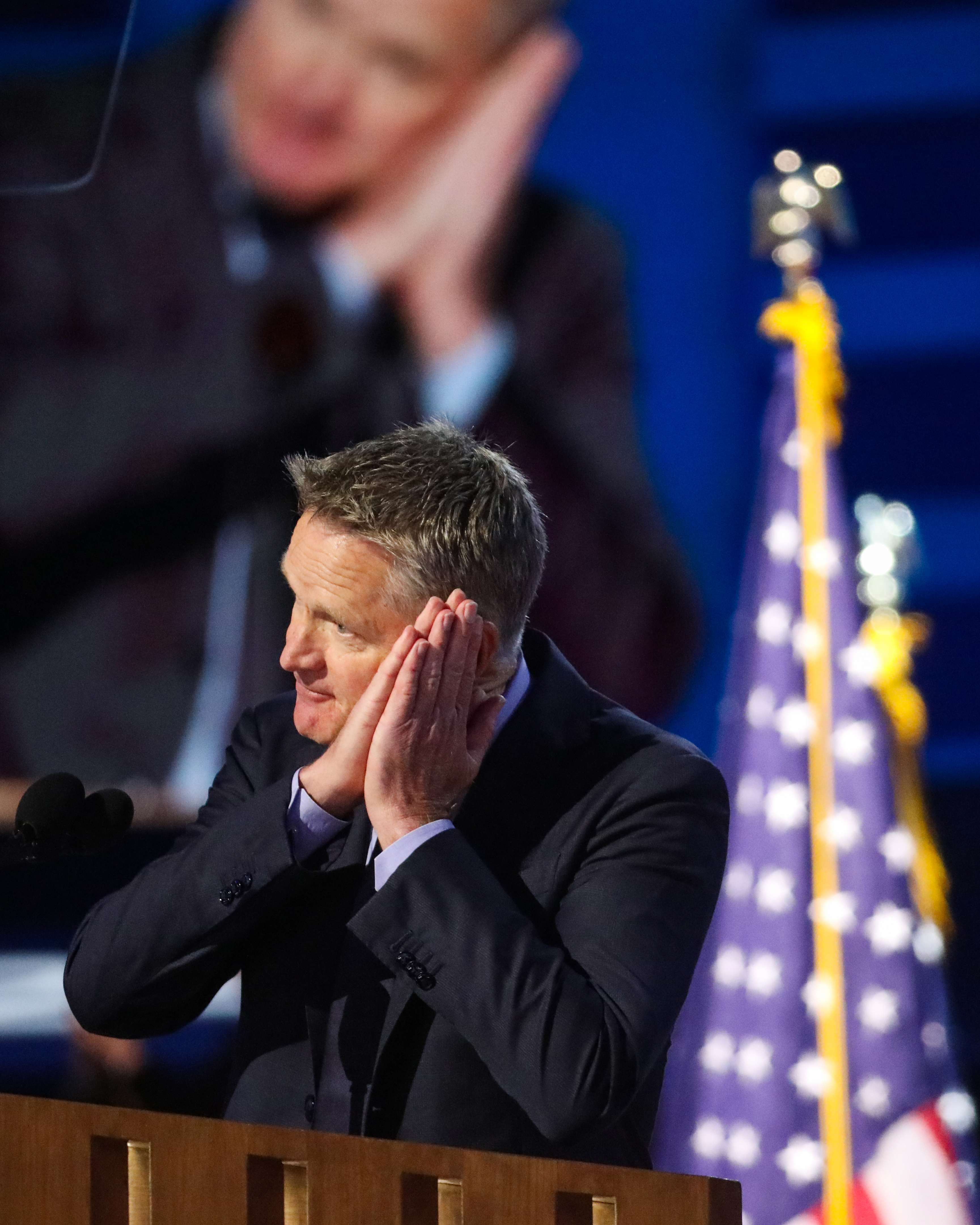 A man at a podium holding his hands to his ears. The American flag is in the background, and a large screen behind him displays the same image
