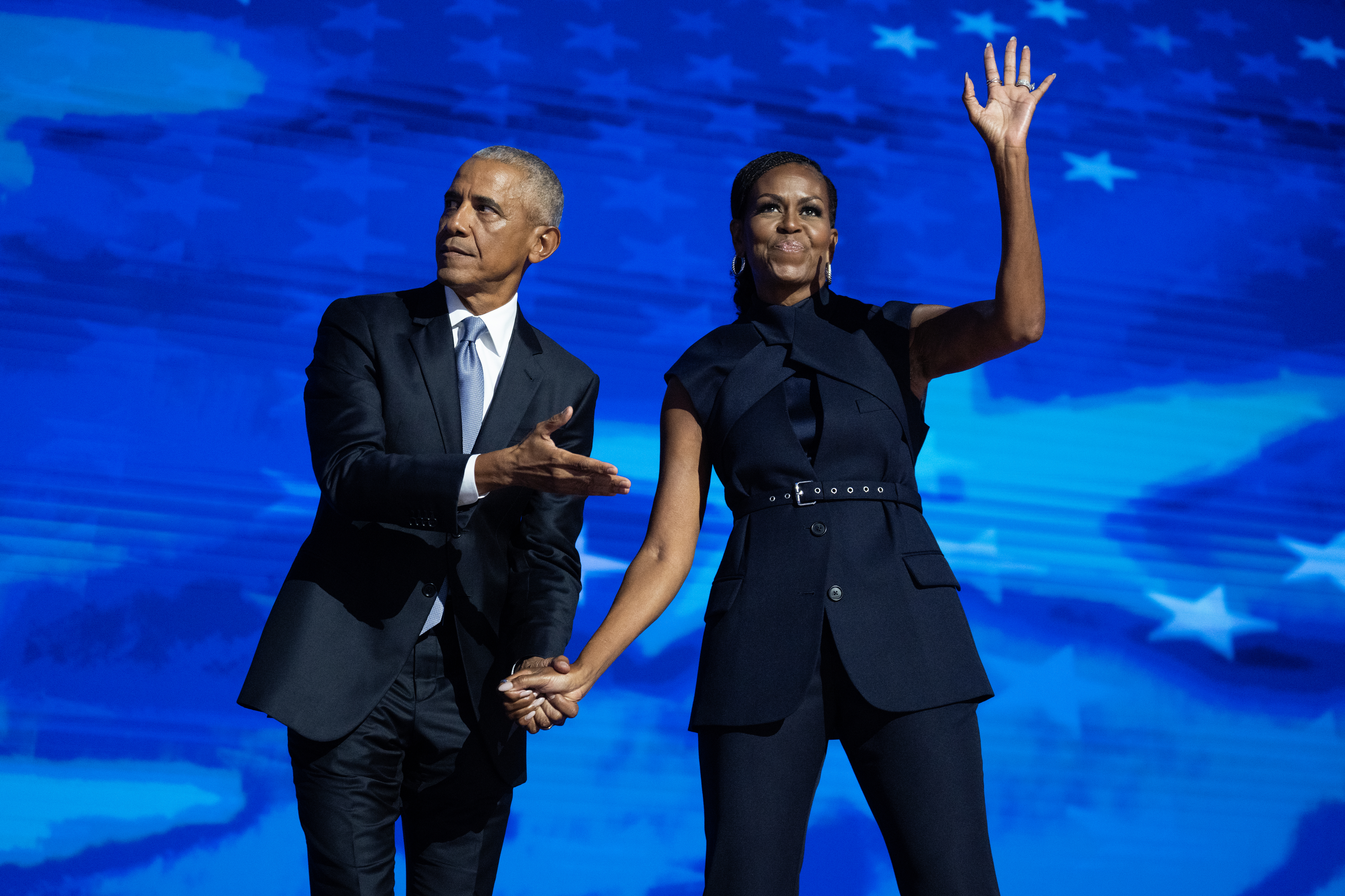Barack Obama and Michelle Obama hold hands on stage, wearing formal black outfits, while Michelle waves to the audience