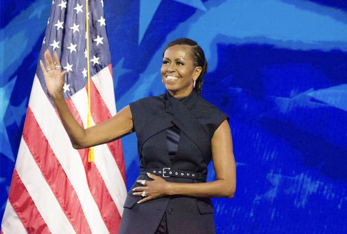 Michelle Obama smiling and waving, standing beside an American flag, wearing a sleeveless, belted black dress with a high neckline