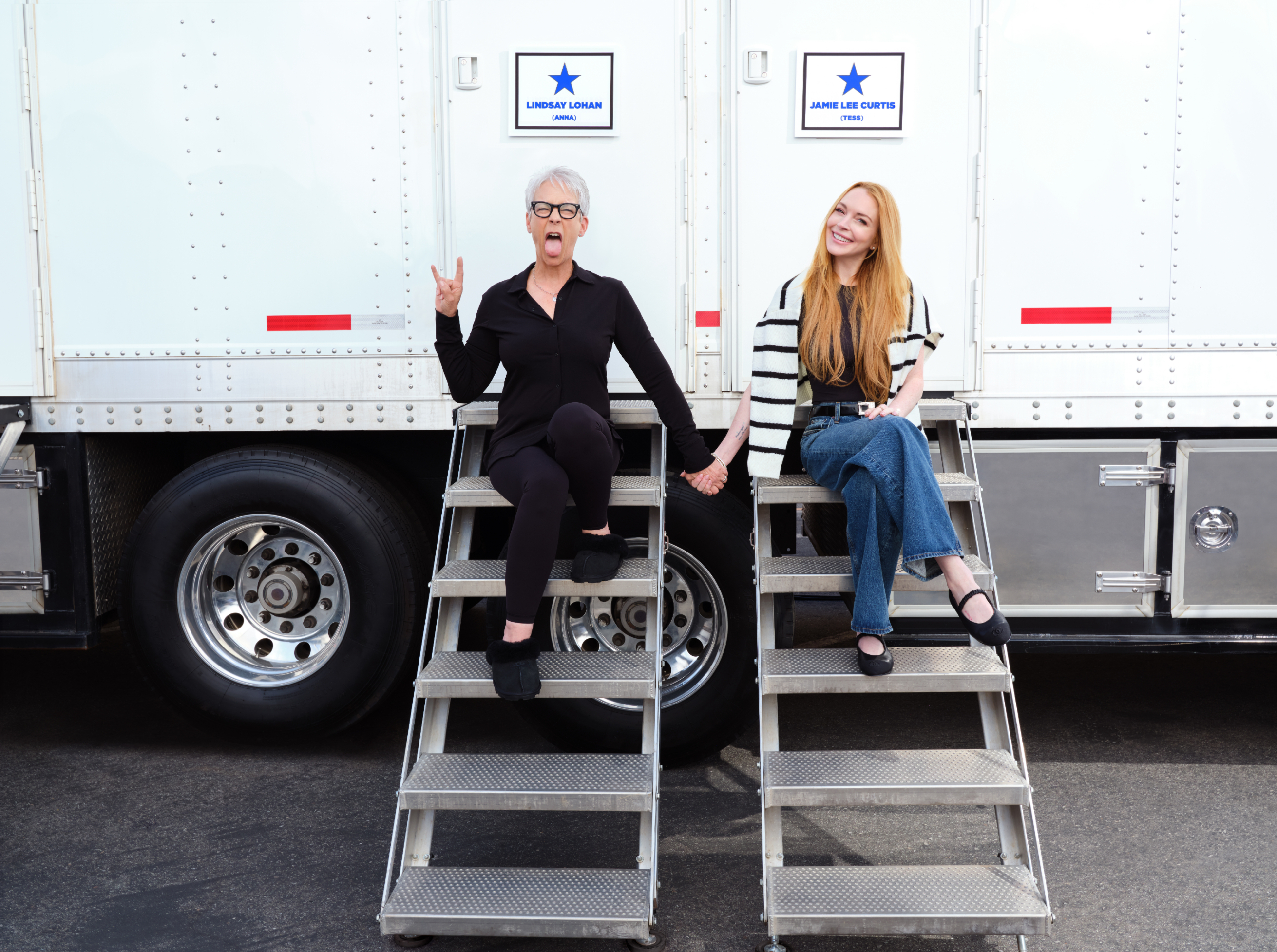 Jamie Lee Curtis (left) and Lindsay Lohan (right) sit on trailer steps. Curtis is wearing a black outfit; Lohan is dressed in a striped top and jeans. They hold hands