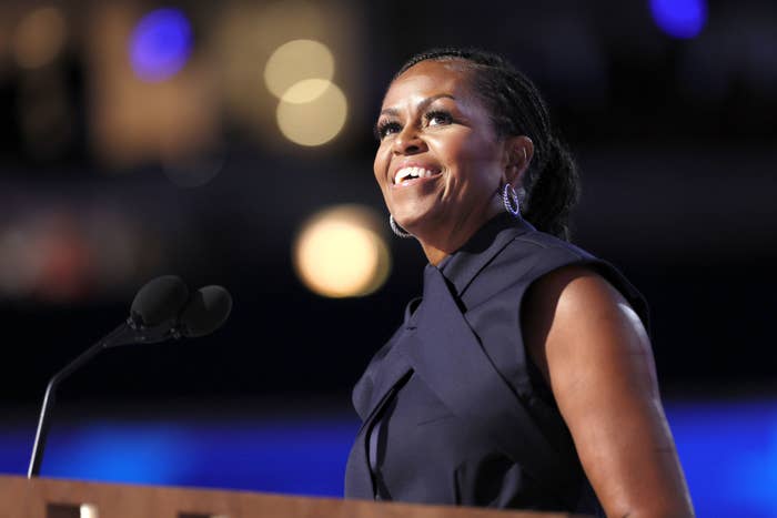 Michelle Obama smiles at a podium, wearing a sleeveless, high-neck dress with a large bow detail, speaking at an event