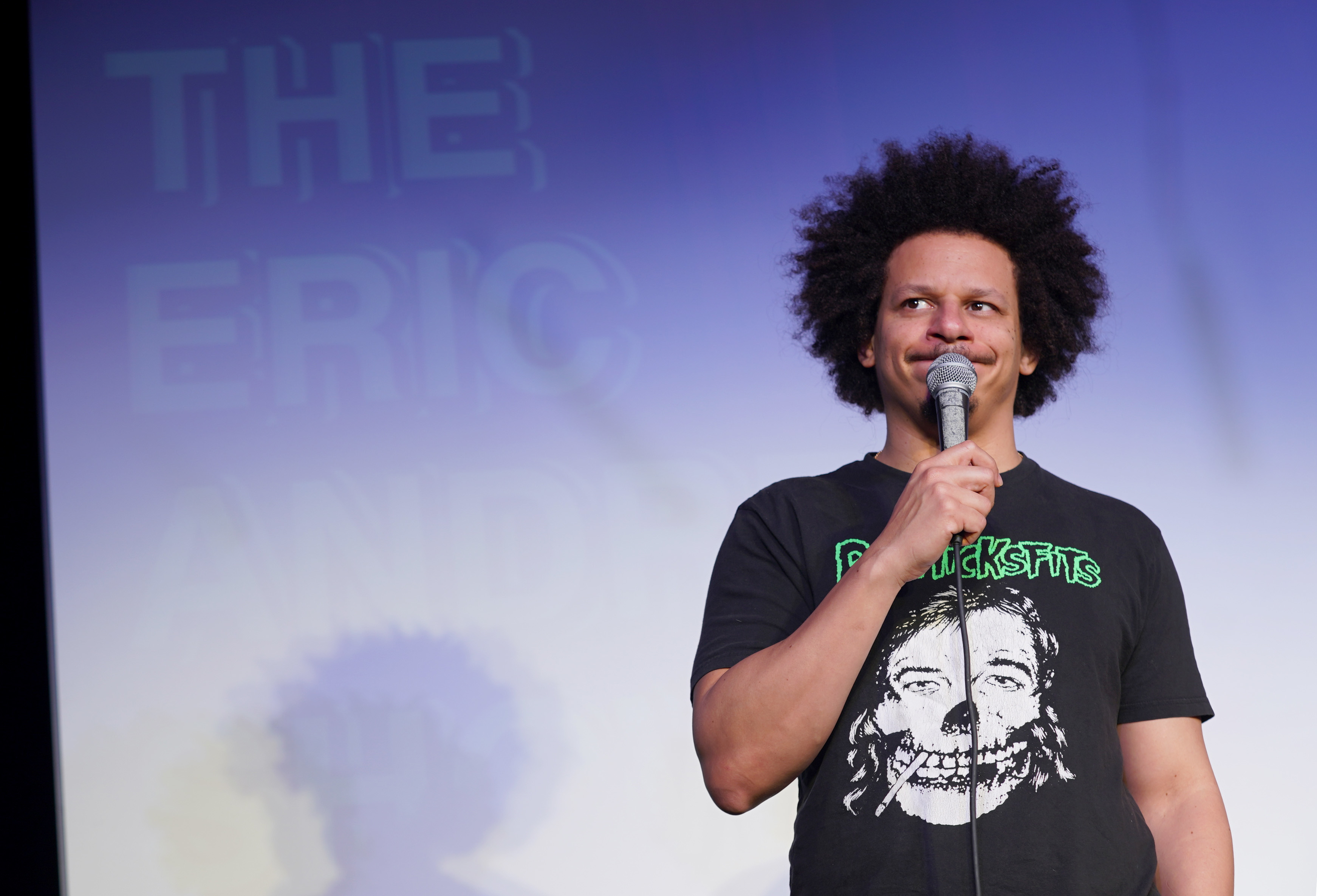 Eric Andre performing stand-up comedy on stage, holding a microphone, wearing a graphic t-shirt with "Dicksfits" printed on it. "The Eric Andre Show" is visible in the background