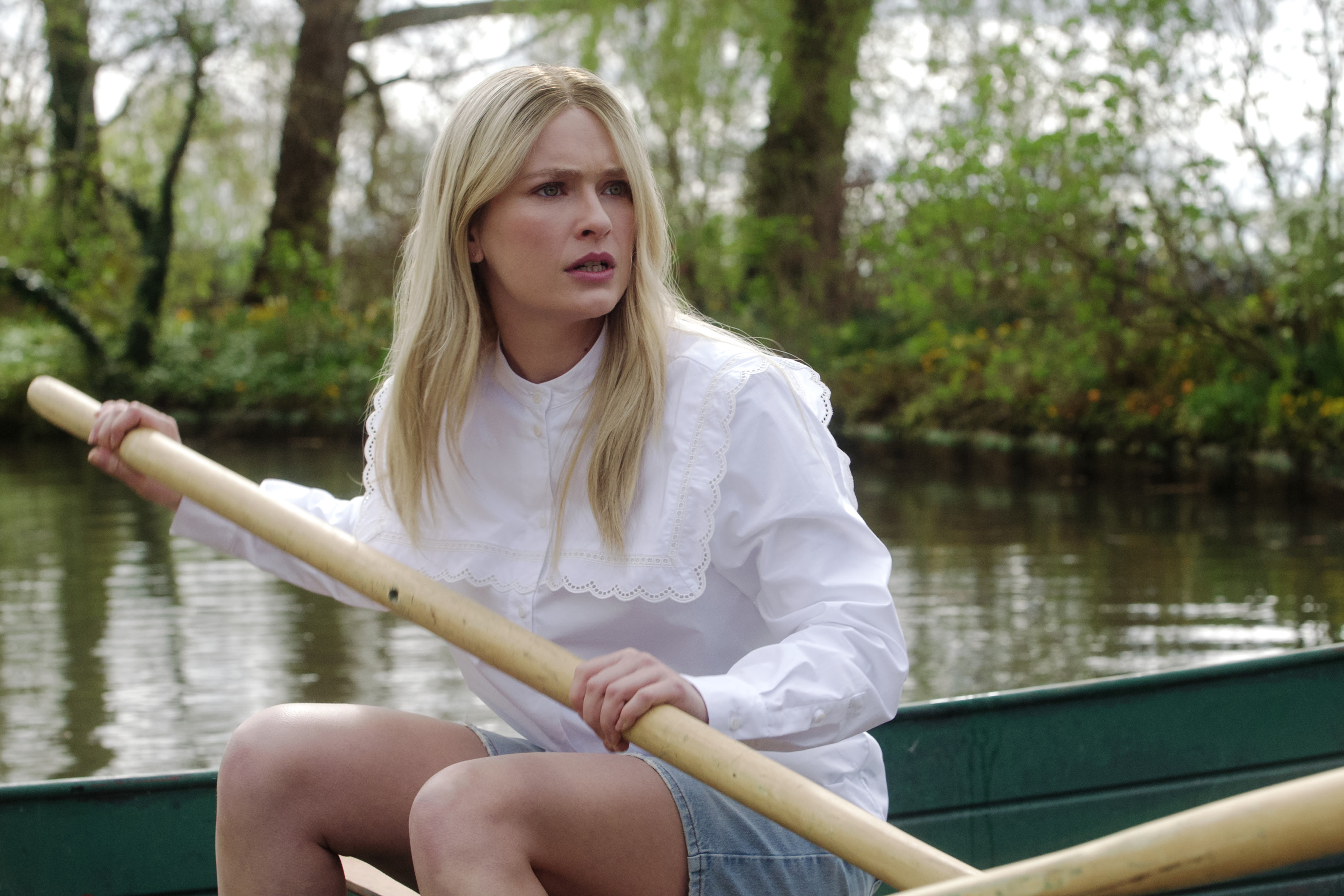 Camille paddling a boat on a calm river, wearing a white blouse with lace detail and denim shorts