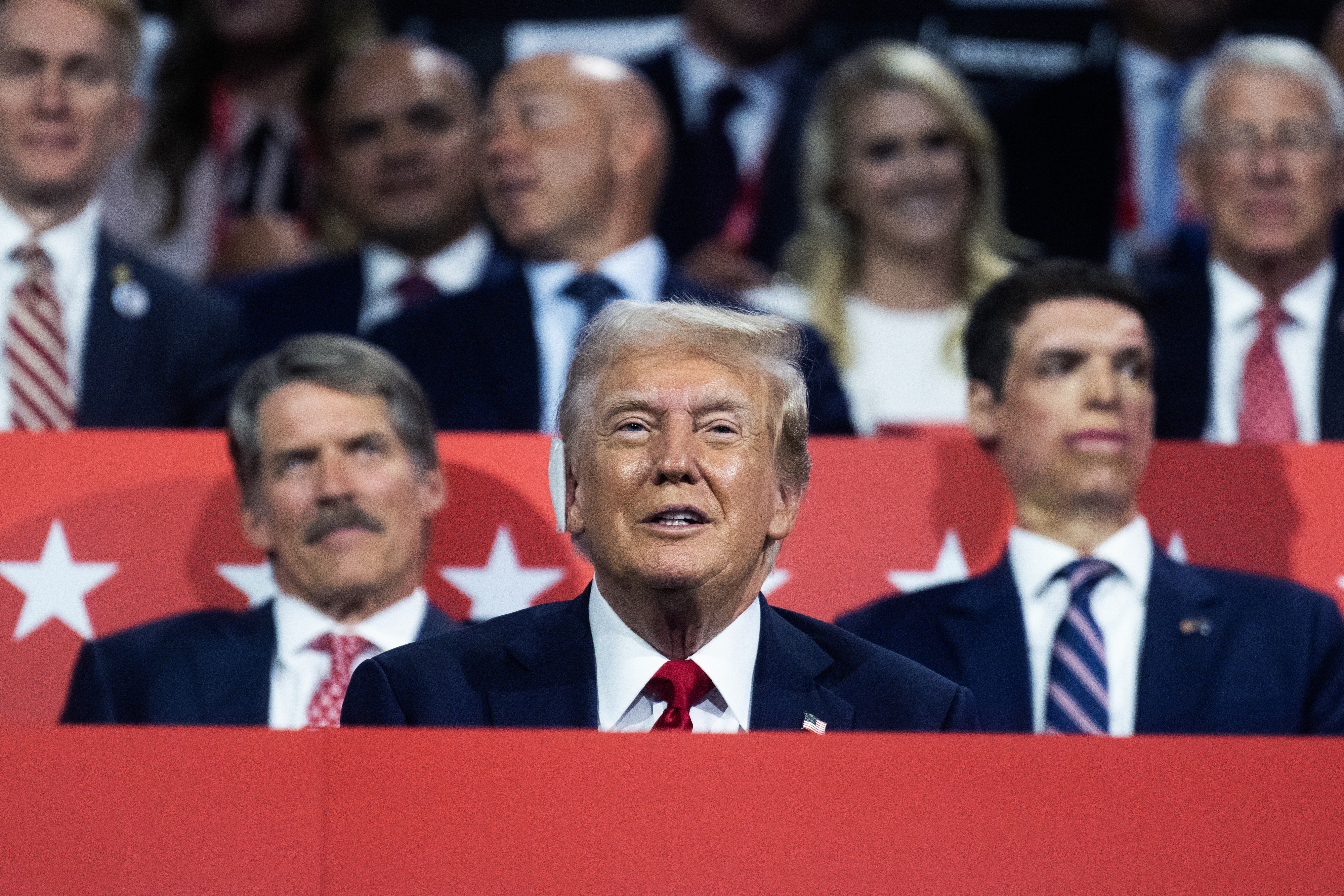 A group of people, including Donald Trump, is seated at a public event. Donald Trump, in the center, is wearing a suit and tie. Other individuals are present in the background