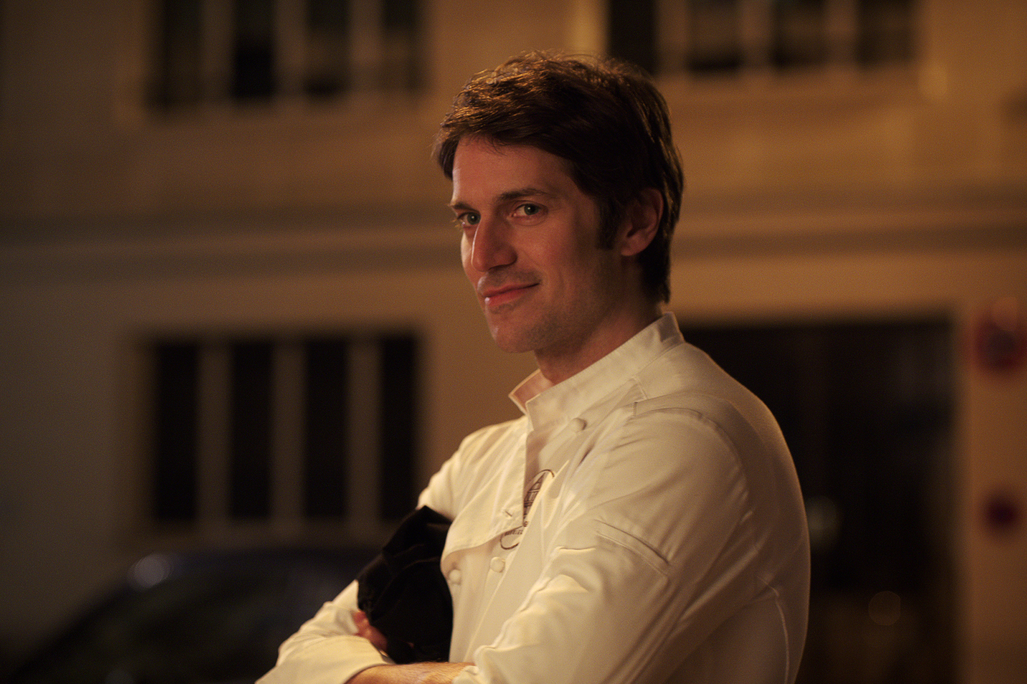 A man, dressed in a white shirt, standing with a slight smile in an urban setting during the night