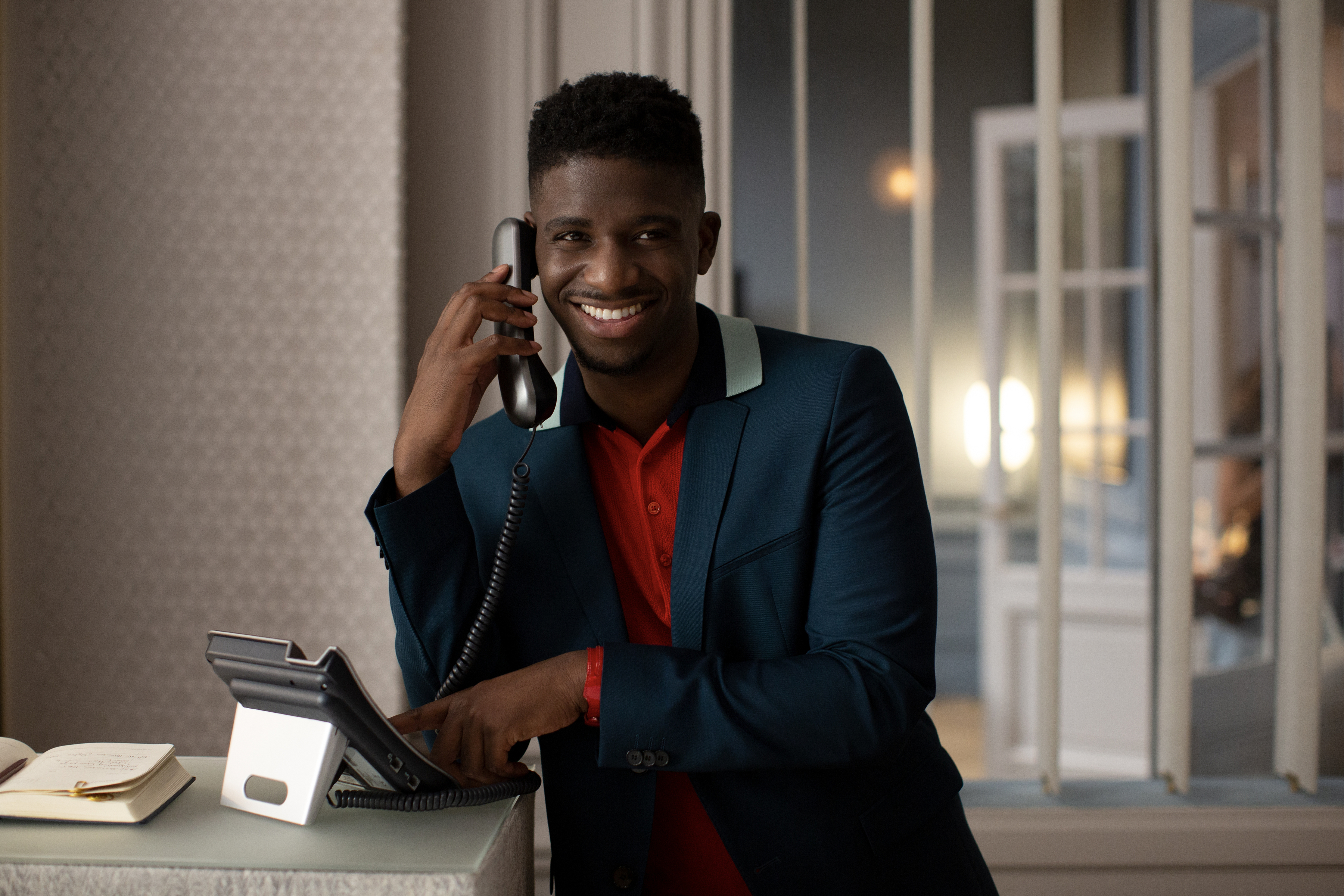 Man in a suit and red shirt smiles while talking on the phone and using a computer. Office setting in the background