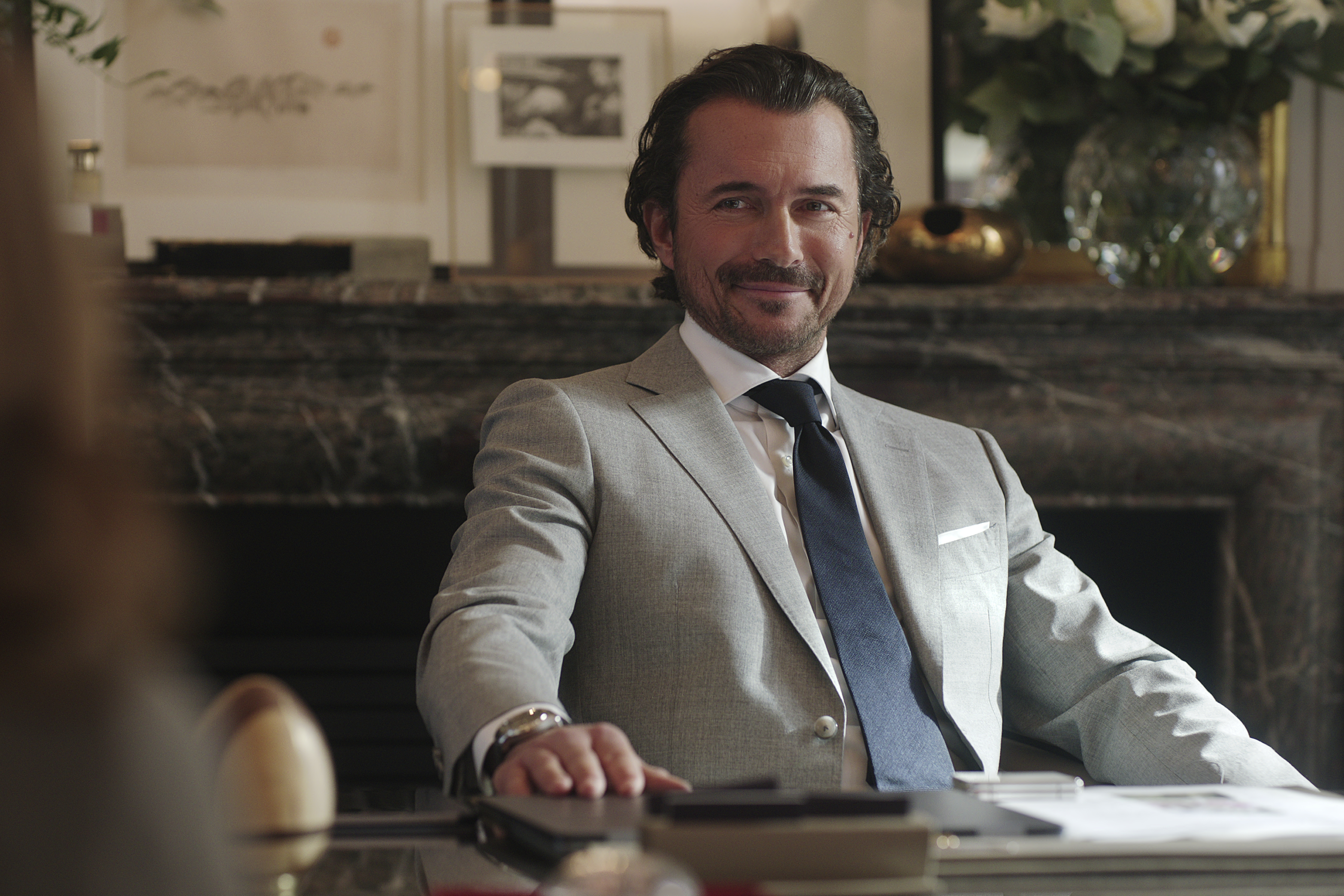 Man in a gray suit with a dark tie, smiling while seated at a desk in a well-decorated office