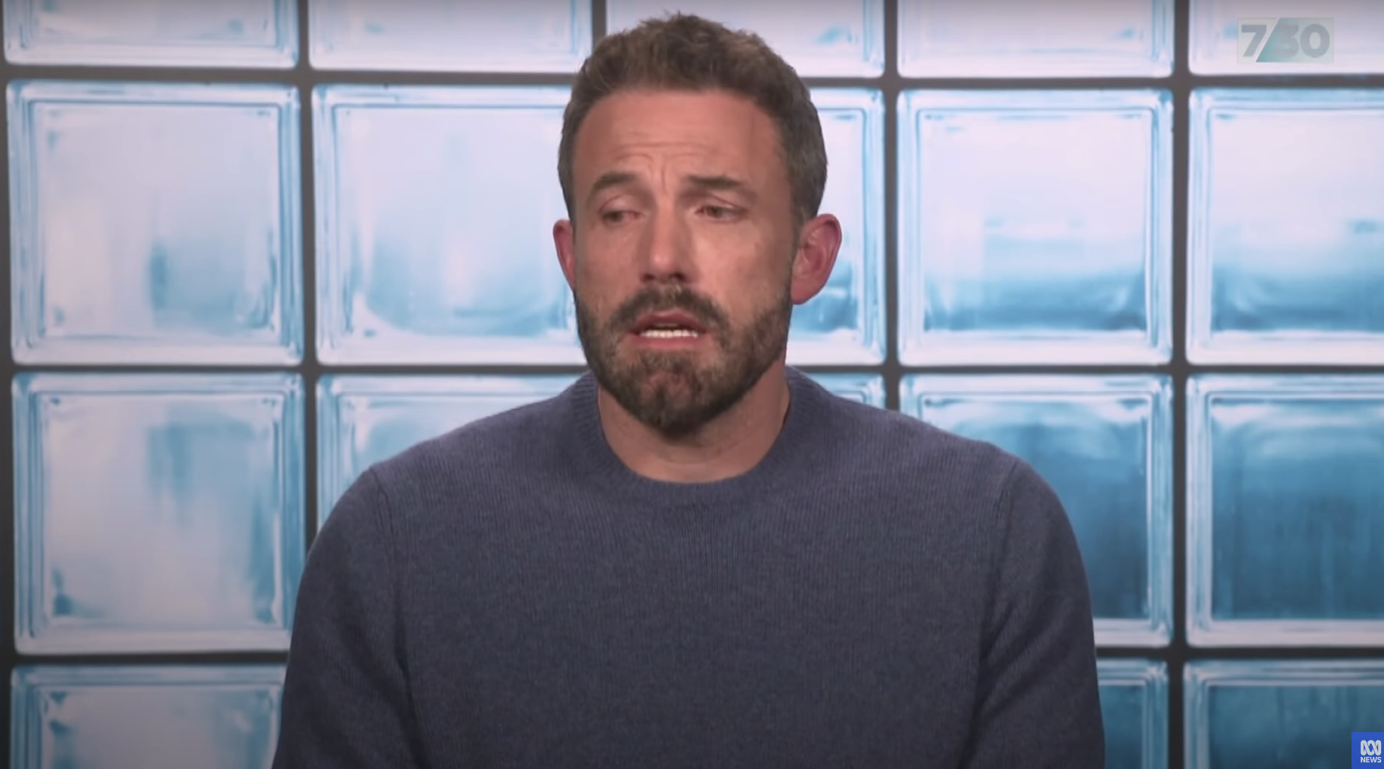 Ben Affleck with short hair and a beard, wearing a sweater, speaks with a serious expression in front of a background of glass blocks