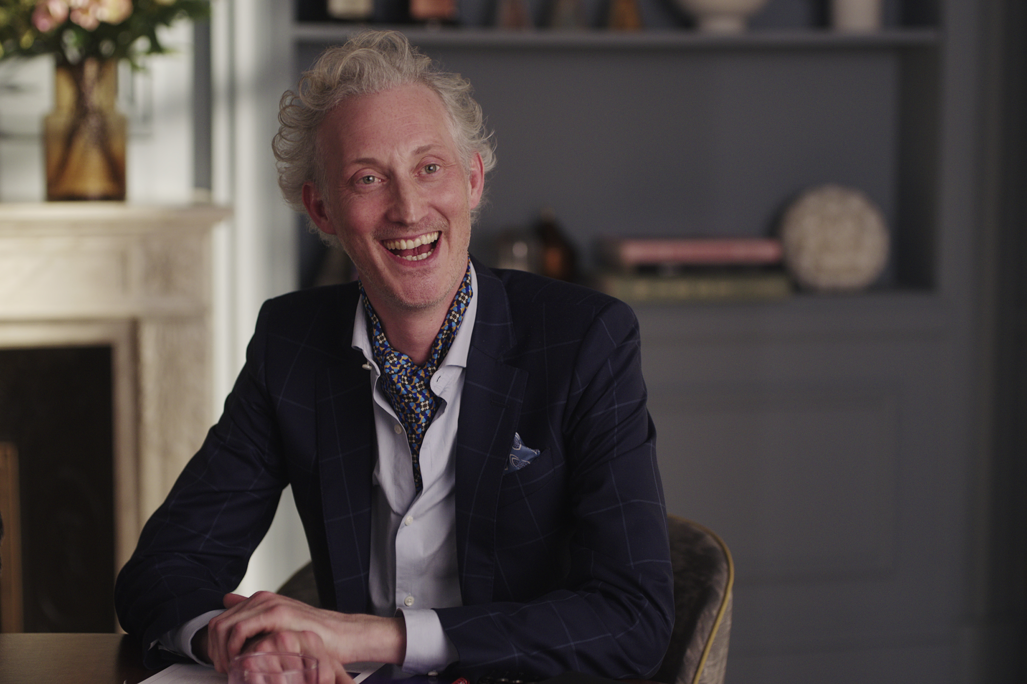 An elegantly dressed man with curly hair, wearing a dark suit and patterned scarf, smiles while seated at a table in an upscale room