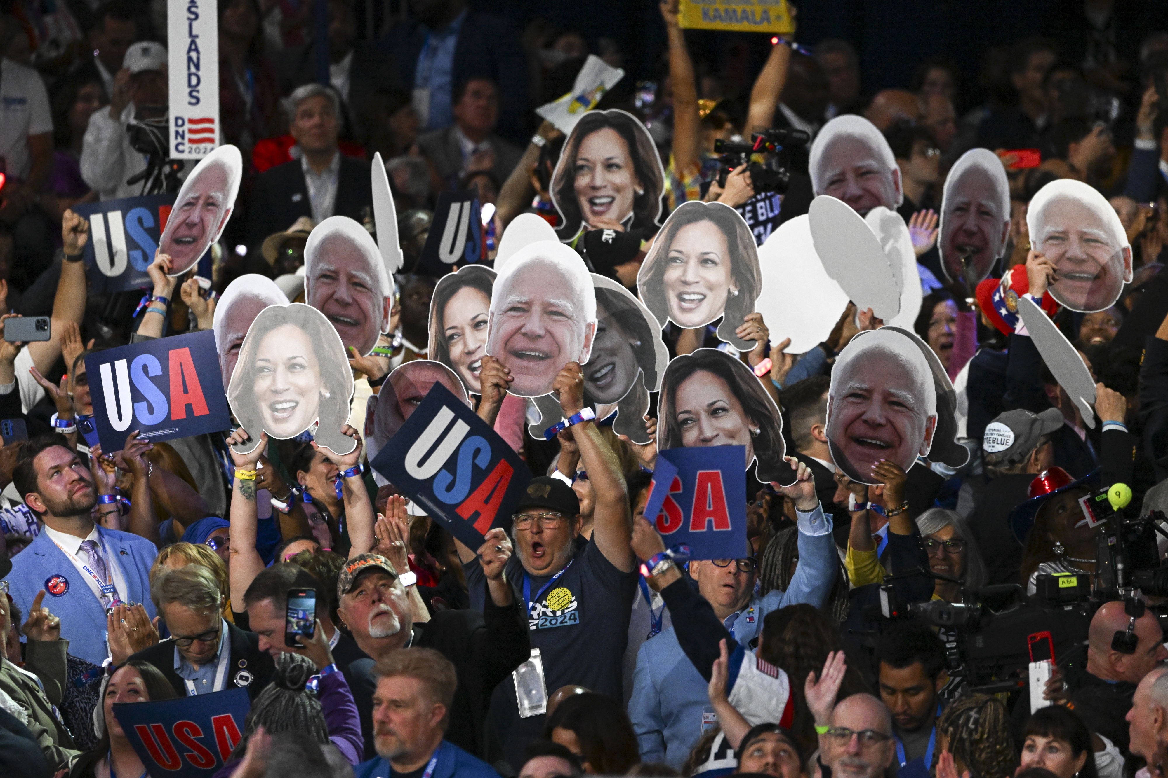 Crowd holding oversized cutouts of Bernie Sanders and Kamala Harris faces, along with USA signs, at a lively event