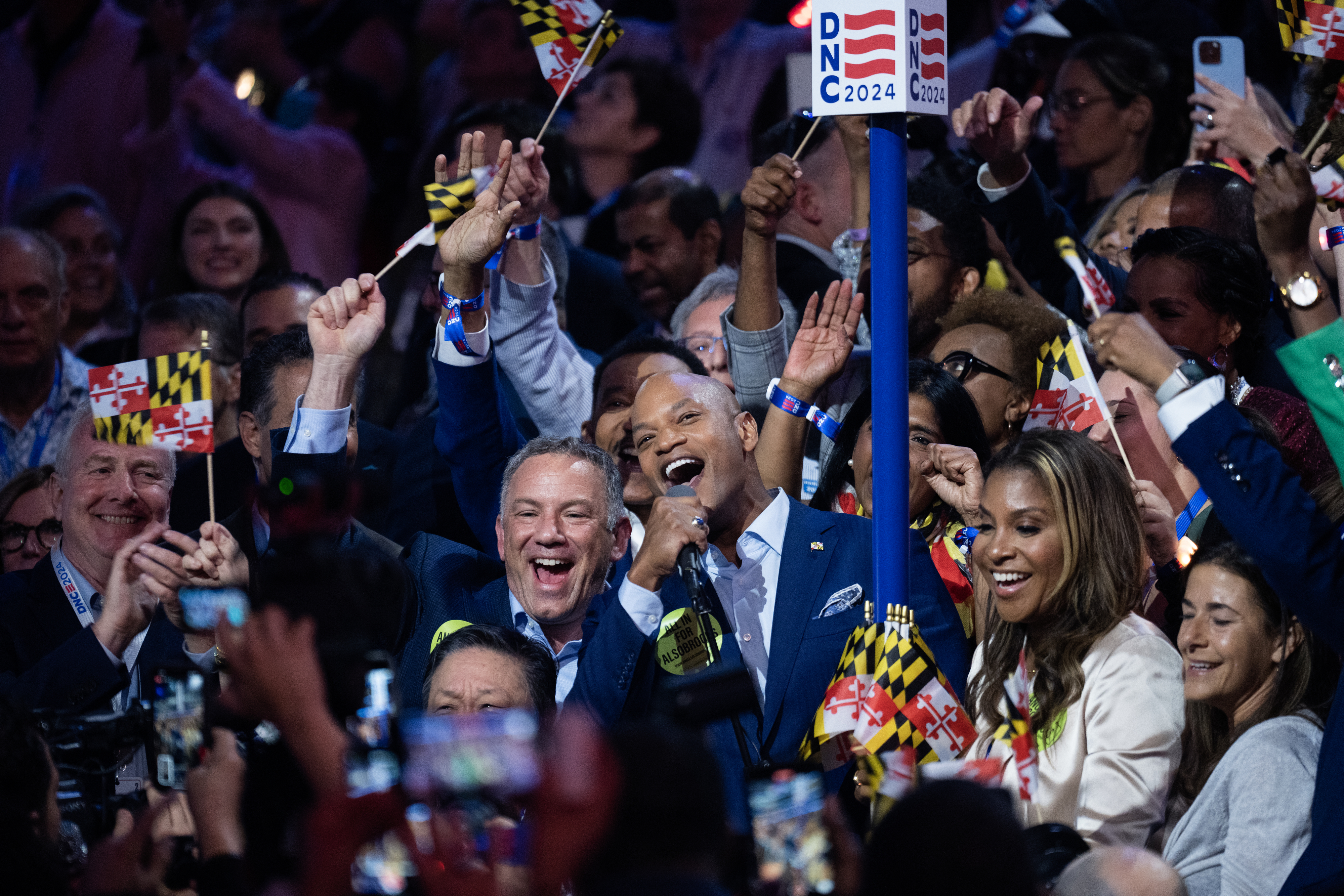 Wes Moore and a group of enthusiastic supporters, holding Maryland state flags, celebrate at a lively event under a sign reading "DNC 2024."