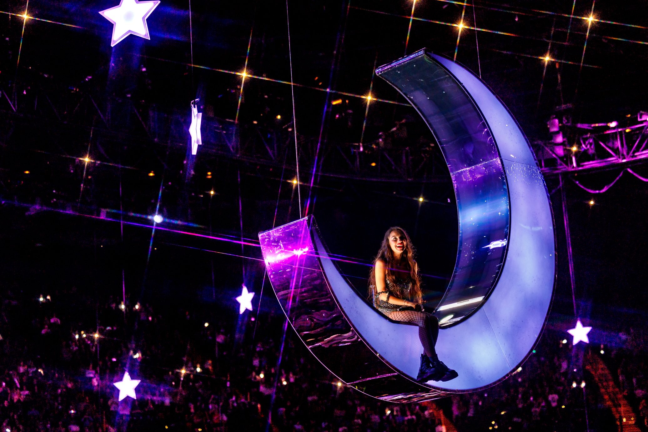 Ariana Grande performs while seated on a crescent moon prop suspended in the air. The background is adorned with stars