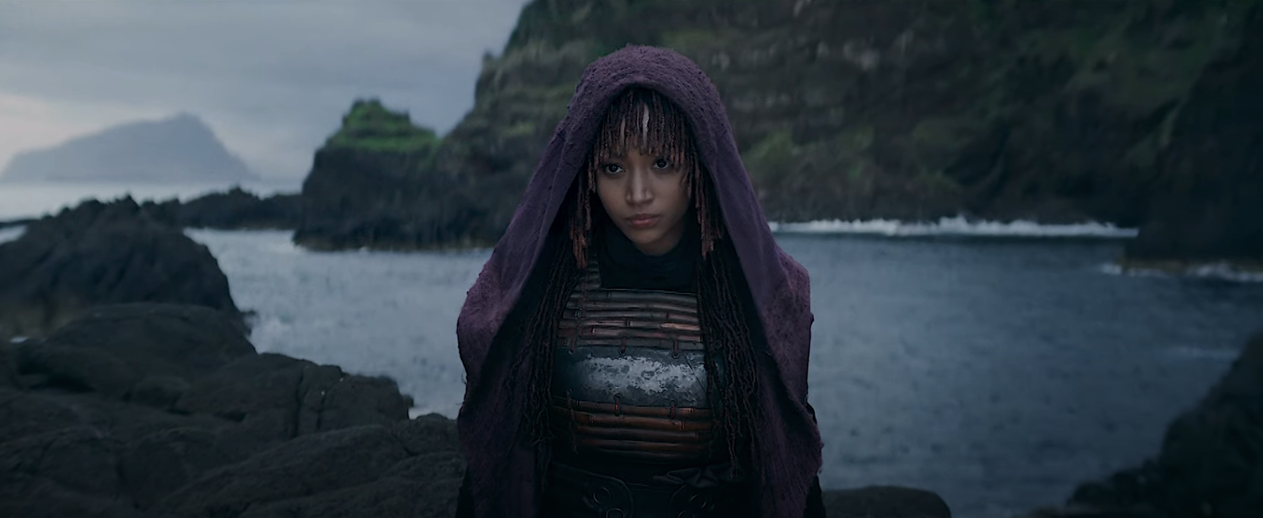 A Sith stands on a rocky coastline in a hooded cloak, staring intently. The scene is moody with ocean waves and cliffs in the background