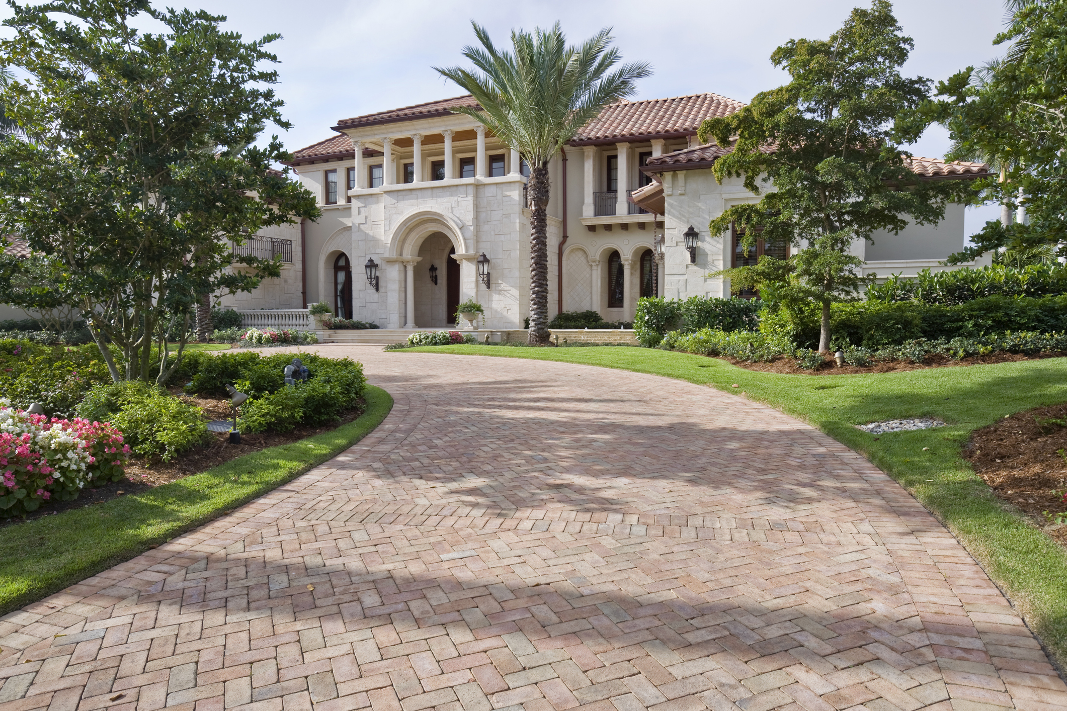 Luxury home with a grand driveway, palm trees, and manicured landscaping. Two stories with a Mediterranean-style design, featuring an arched entrance
