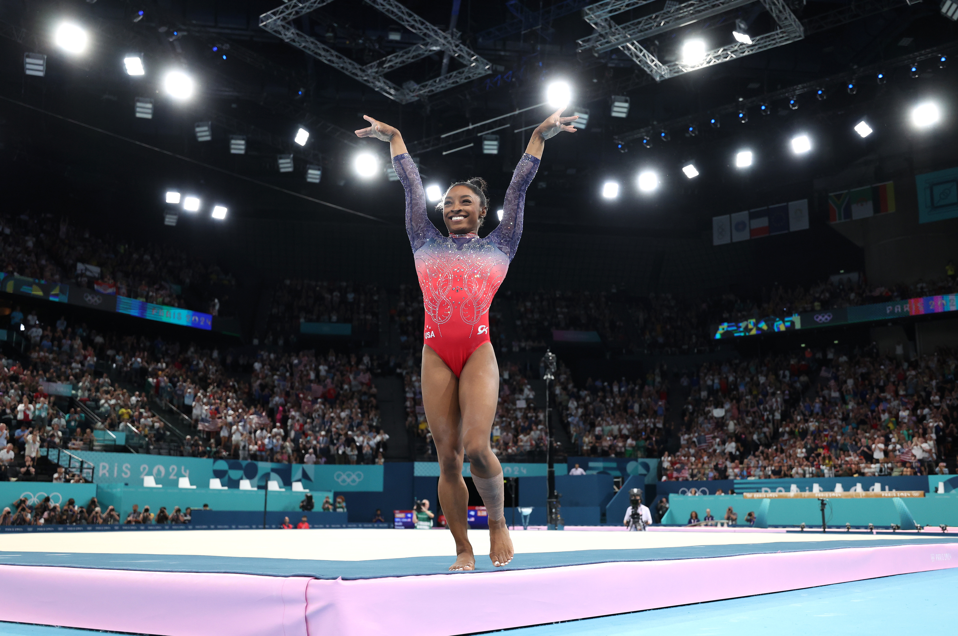 Simone Biles stands on a gymnastics mat with her arms raised in victory at the 2021 Olympic Games, surrounded by cheering fans in a stadium