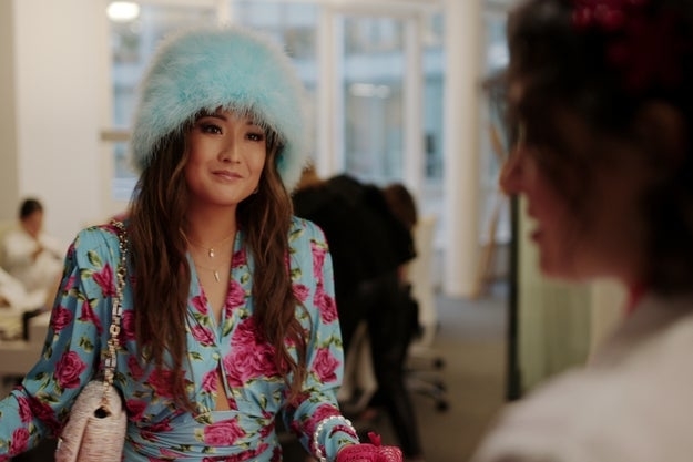 Ashley Park, dressed in a floral outfit with a fuzzy hat, talks to Lily Collins' character in a stylish office setting