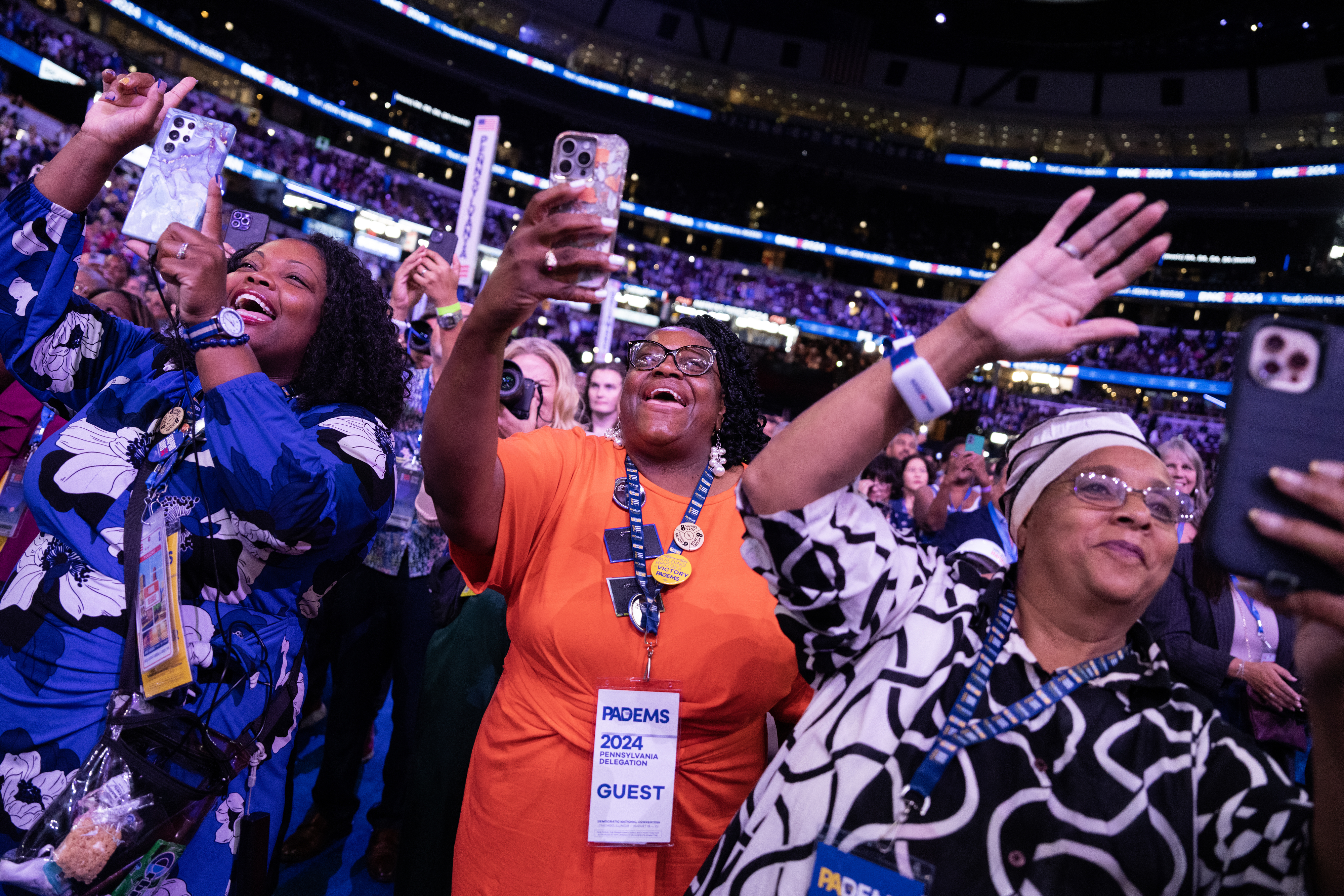Three women holding phones excitedly capture moments at an event, with visible crowd and event signage reading "PDCEMS 2024" and "GUEST."