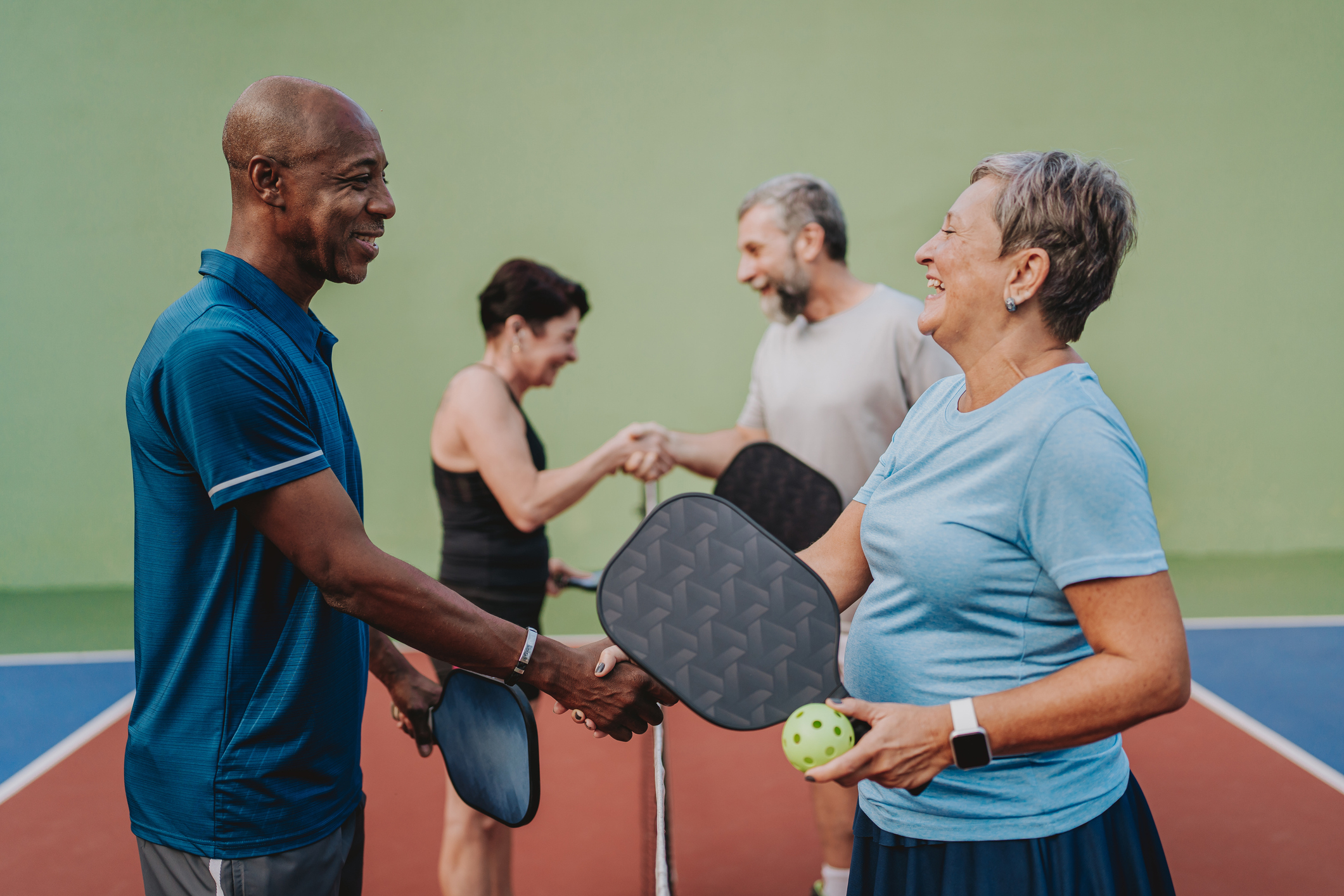 An older couple plays pickleball on a court alongside another couple. They smile and shake hands, sharing good sportsmanship and camaraderie