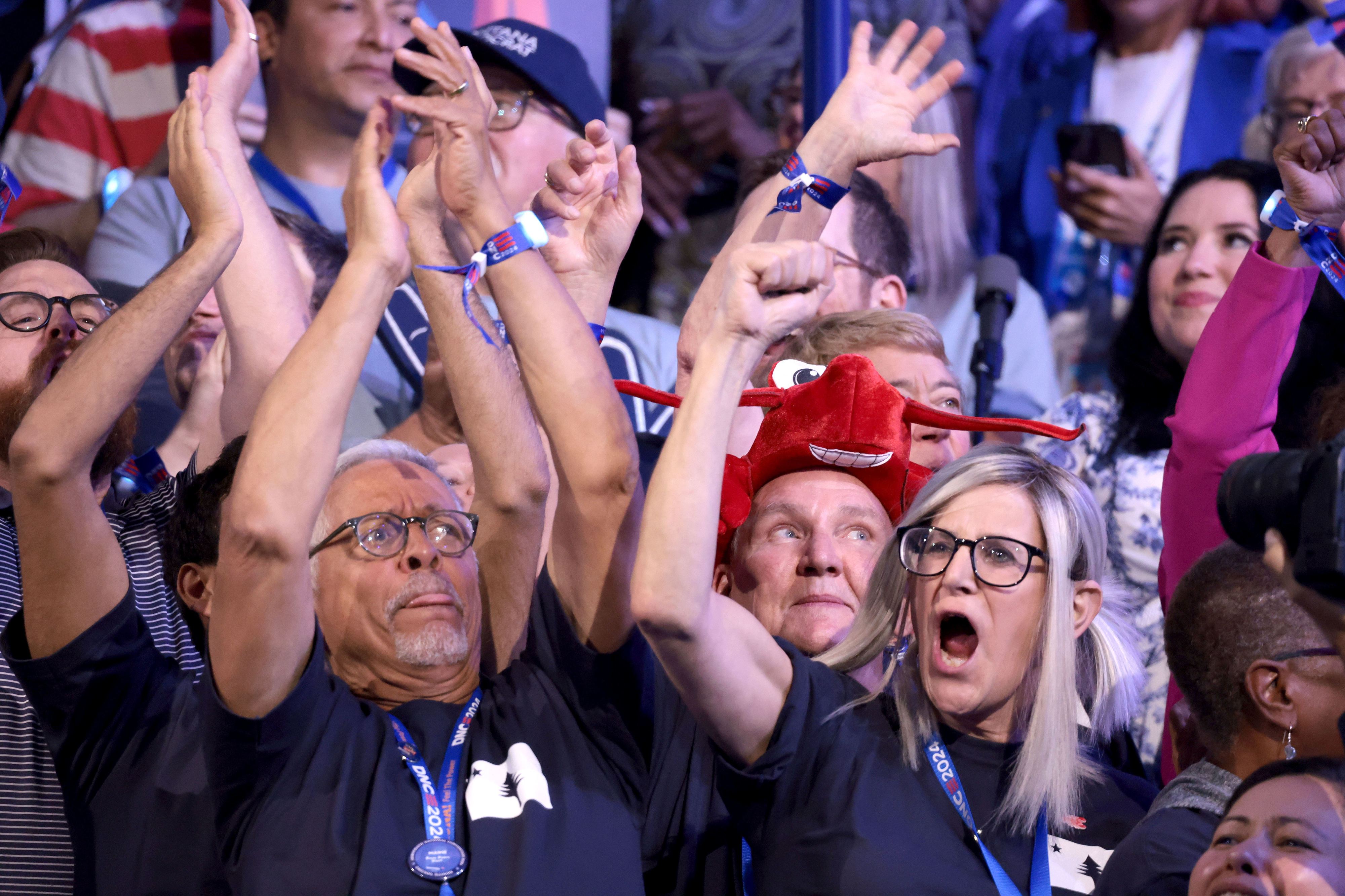 A crowd at an event where attendees, wearing blue lanyards and various accessories, are enthusiastically raising their hands and cheering